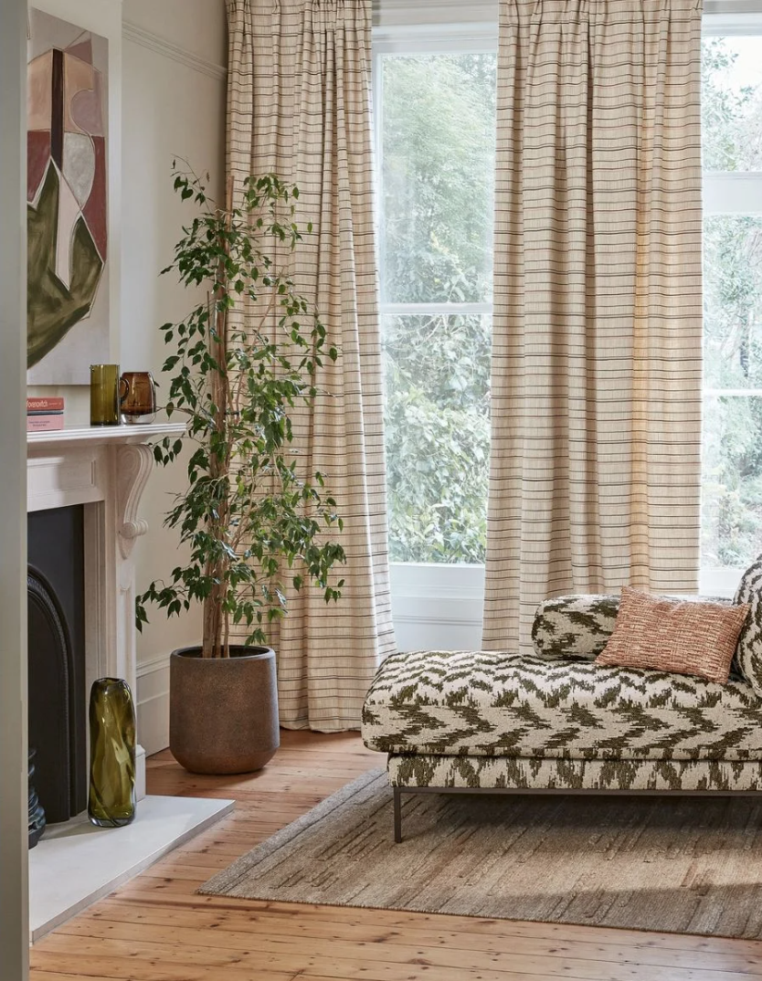 A cozy living room corner featuring a patterned sofa with pillows, large windows with beige striped curtains, a tall potted plant, a white fireplace mantel with decorative vases, artwork on the wall, and a wooden floor with a patterned rug.
