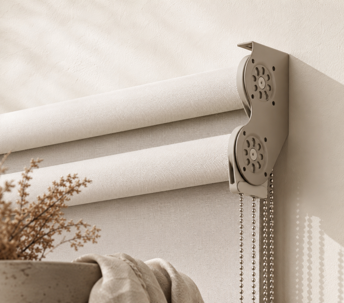 Close-up of a beige window roller blind with metal chain pulls, mounted on a light-colored wall, with a part of a petal-shaped window shade and a flower arrangement visible in the foreground.