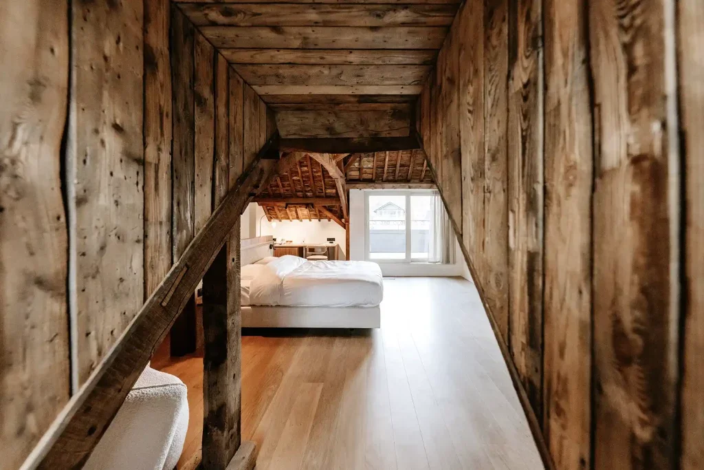 Chambre dans un loft avec mobilier en bois, lit avec draps blancs, grande fenêtre avec vue sur un bâtiment extérieur.