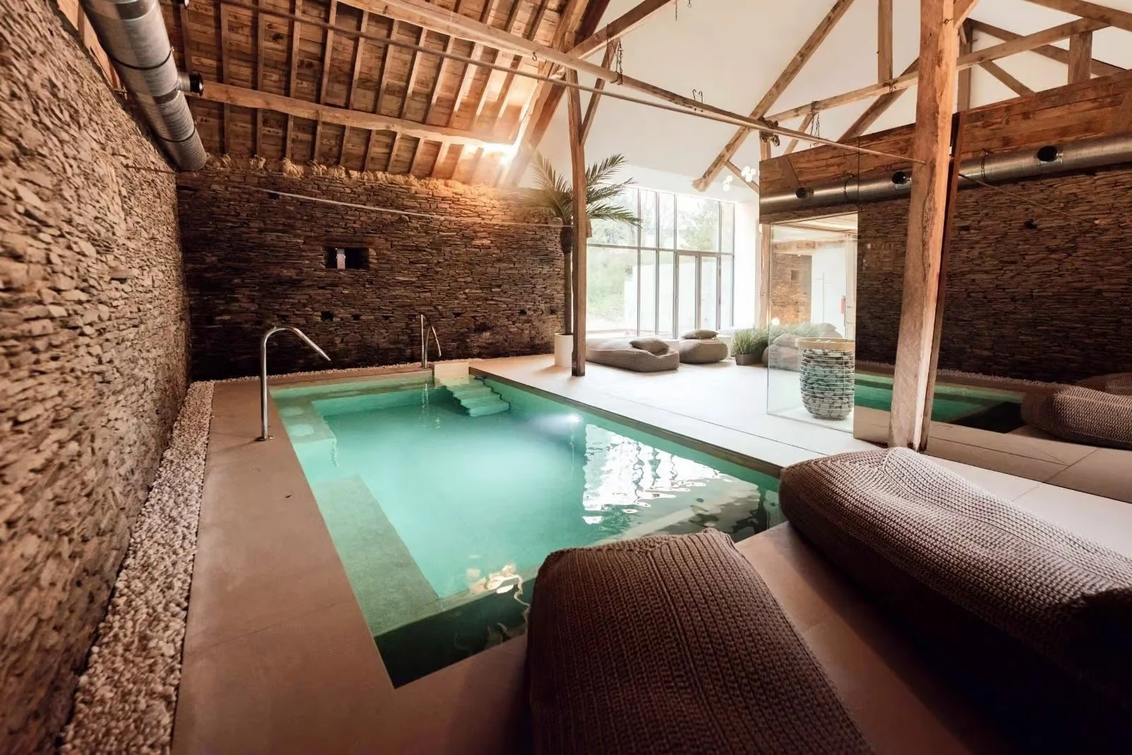 Piscine intérieure avec murs en pierre, plafond en bois, et grand window laissant entrer la lumière naturelle. Coin détente avec coussins et plantes.