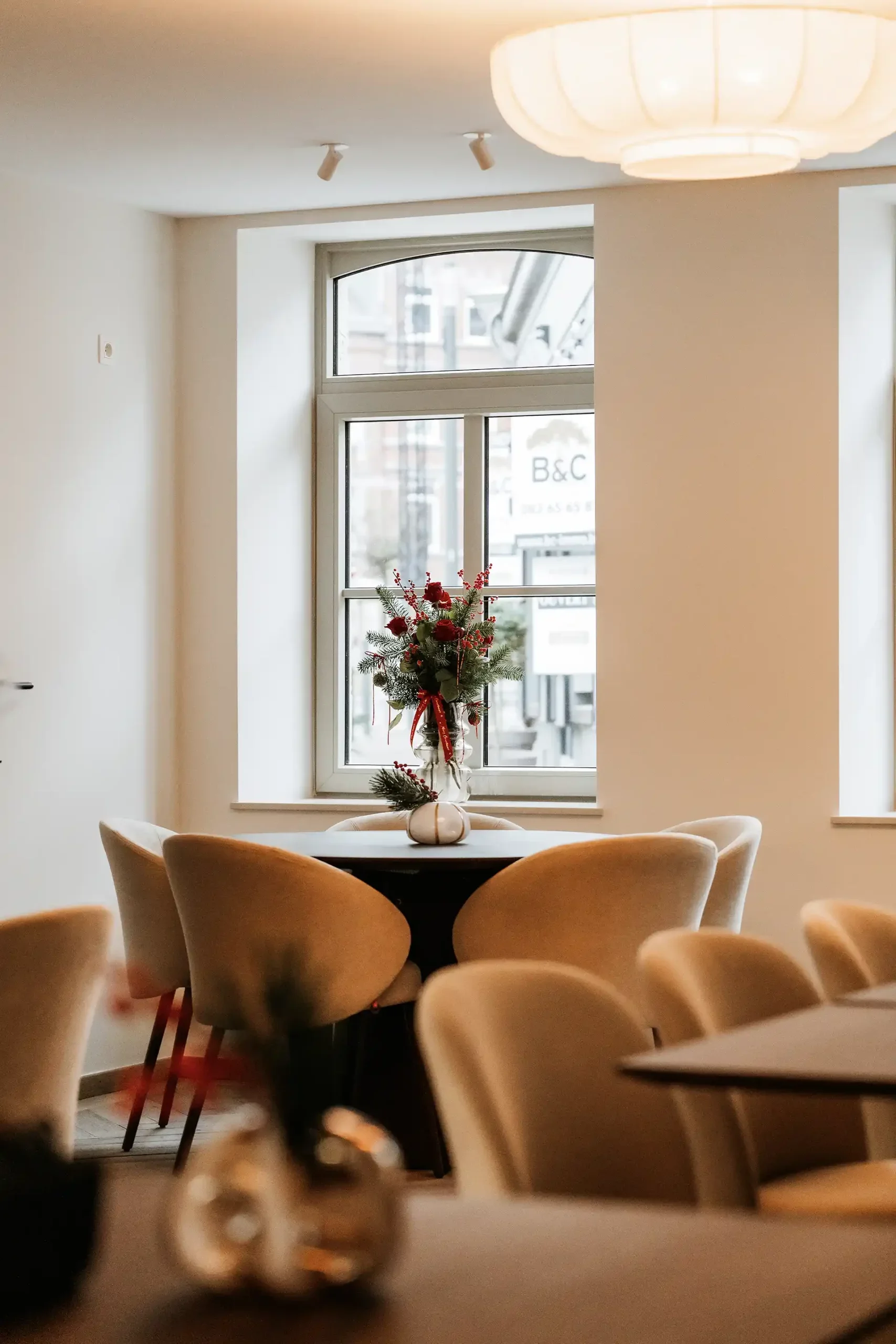 Intérieur d'une salle à manger avec une table ronde, des chaises en tissu beige, et une décoration florale au centre de la table. Une fenêtre laisse passer la lumière naturelle, et un éclairage suspendu au plafond est visible.