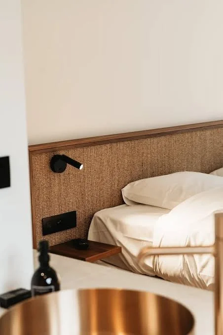 Lits non faits dans une chambre avec une tête de lit en tissu, une lampe de lecture noire, et une petite table de nuit en bois.