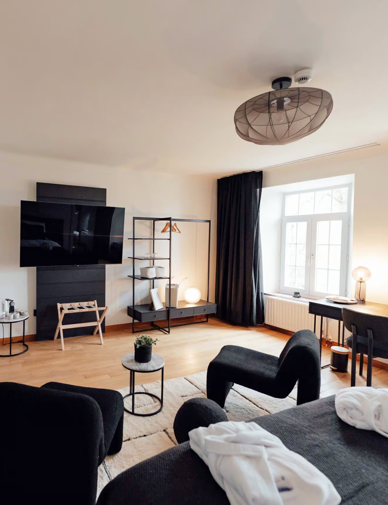 Salon moderne avec canapé noir, petits tables, télévision, étagère noire, grande fenêtre avec rideaux noirs, lampe à papier, décoration sobre, parquet en bois clair.