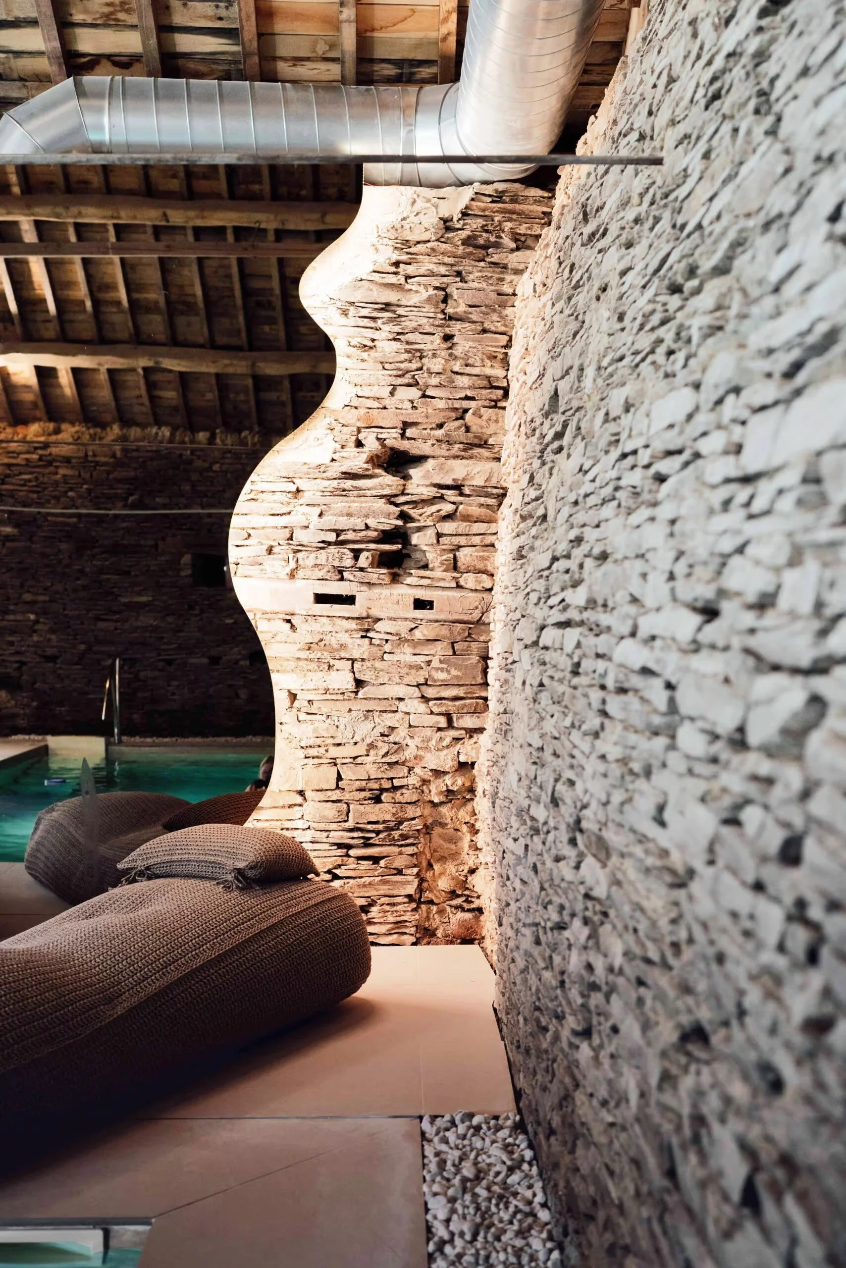 Intérieur d'un espace avec un mur en pierres, un plafond en bois avec poutres apparentes, des coussins sur un sol en pierre, une cheminée et un conduit en métal.