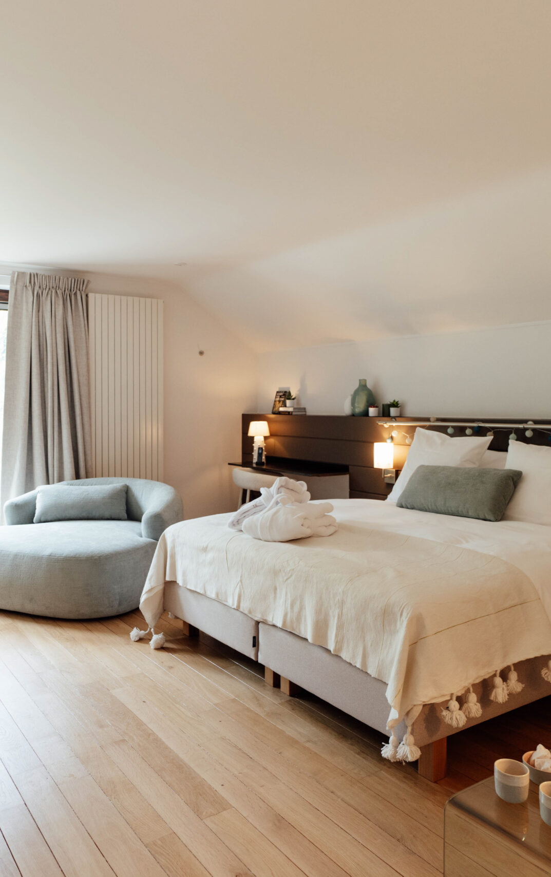 Chambre d'hôtel élégante avec lit fait, canapé gris près de la fenêtre, décor minimaliste et ambiance chaleureuse.