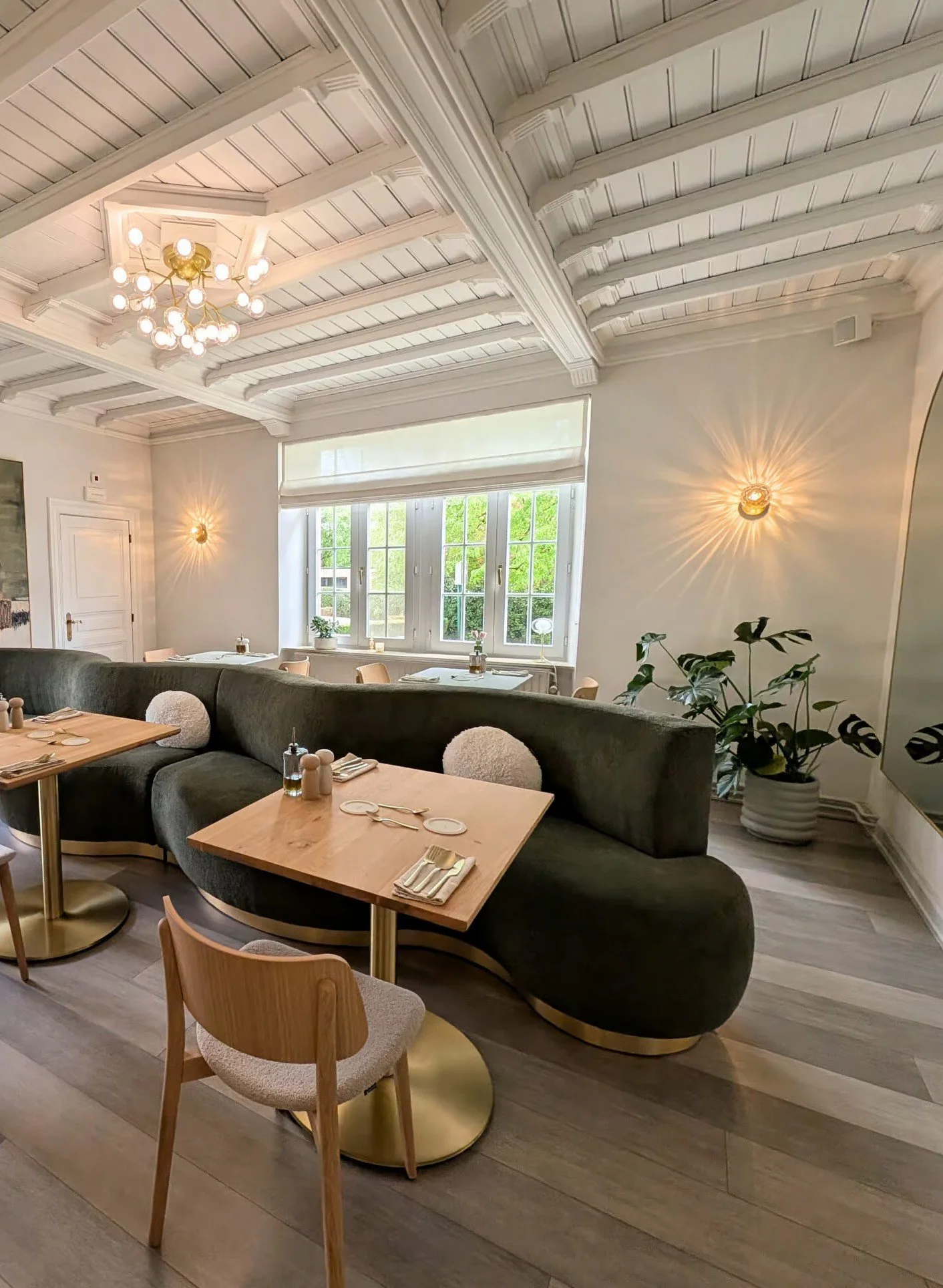 Salle à manger élégante avec un plafond en bois peint en blanc, un lustre moderne, des murs blancs, un grand fenêtre laissant entrer la lumière naturelle, un canapé gris foncé, des tables en bois avec vaisselle, des chaises en bois, et un grand pot avec une plante verte.