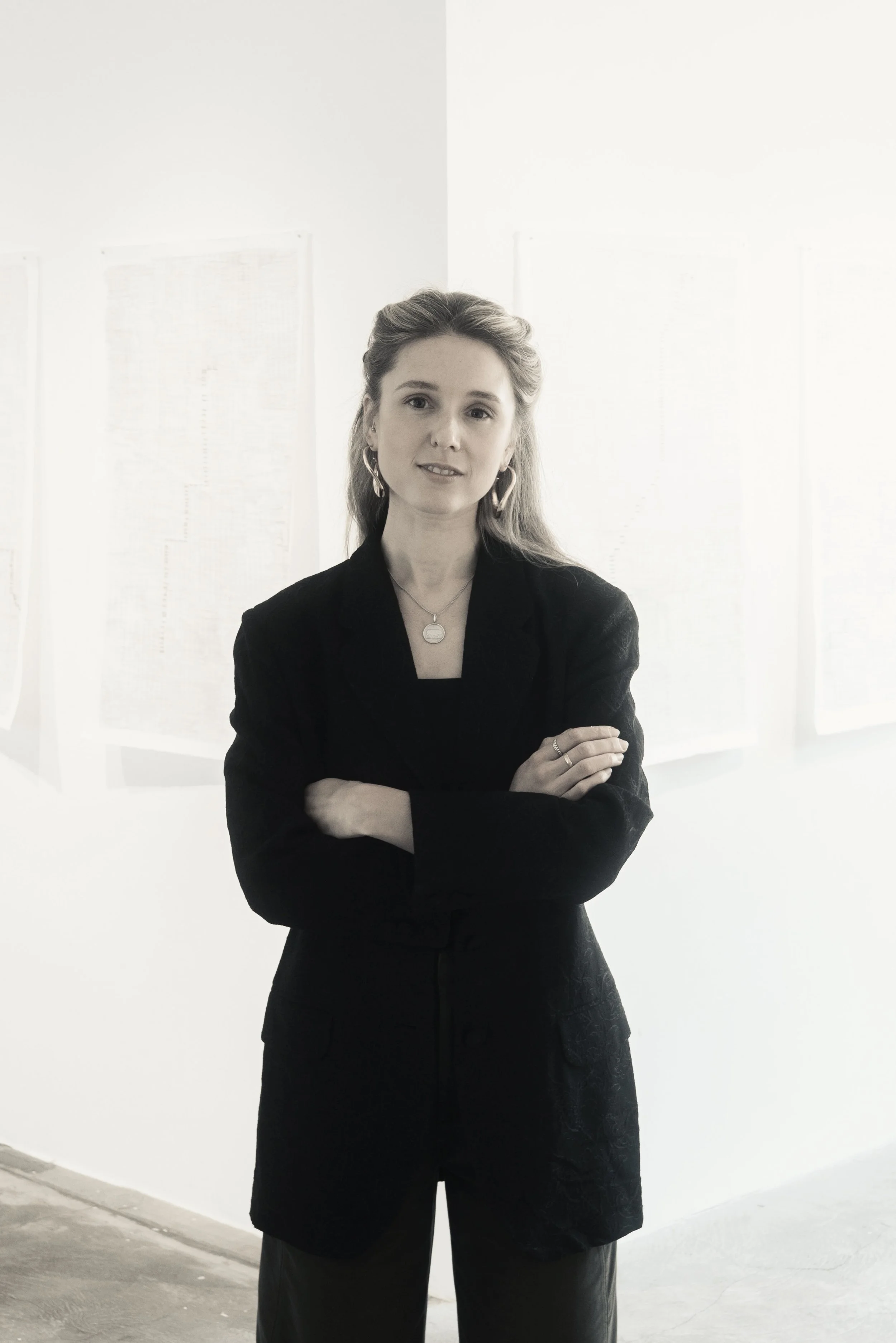 A woman with long, wavy hair, wearing a black blazer and jewelry, standing with arms crossed in front of a white wall with charts or papers, in a bright, minimalistic room.