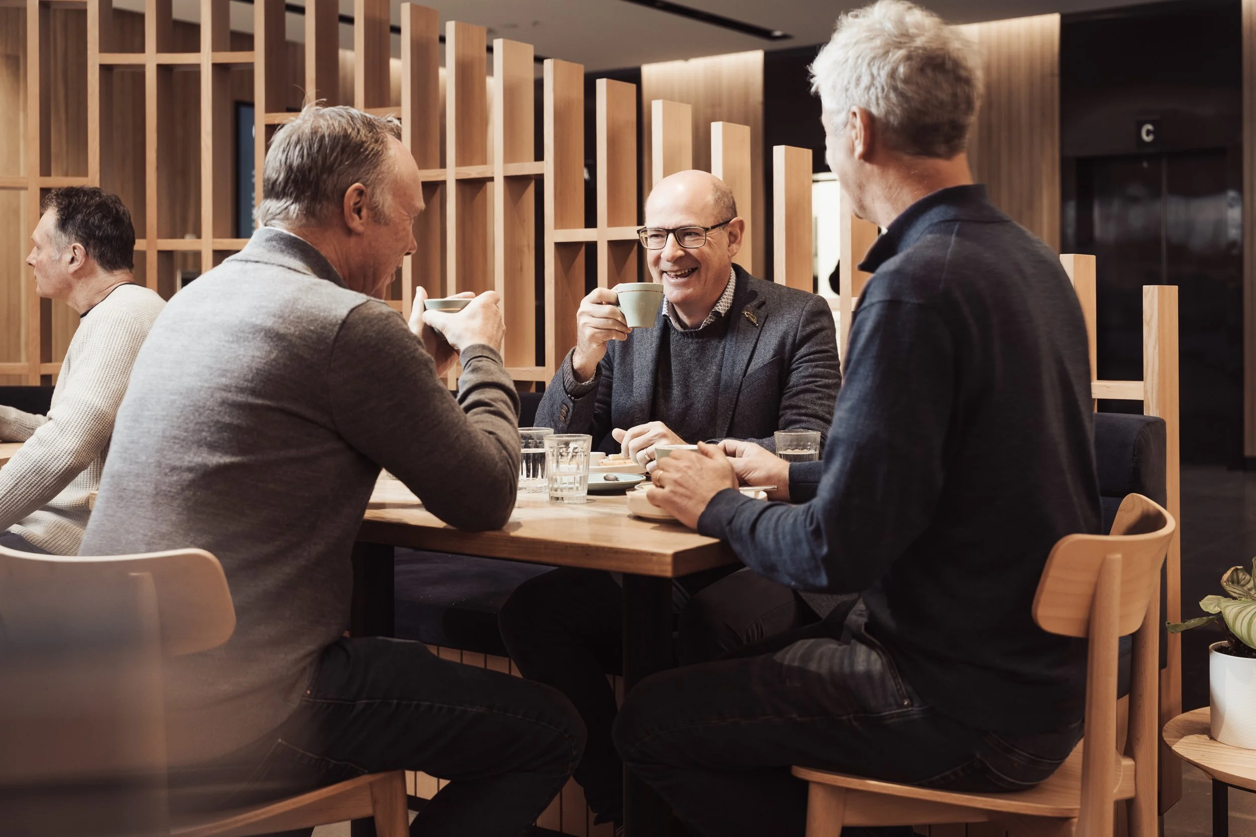 Four men sit at a wooden table in a modern cafe, happily sharing coffee and conversation. One man is smiling broadly while holding a mug, with two others also holding mugs, and the fourth man is turned away from the camera, in a gray blazer. The background features wooden lattice wall partitions and warm lighting.