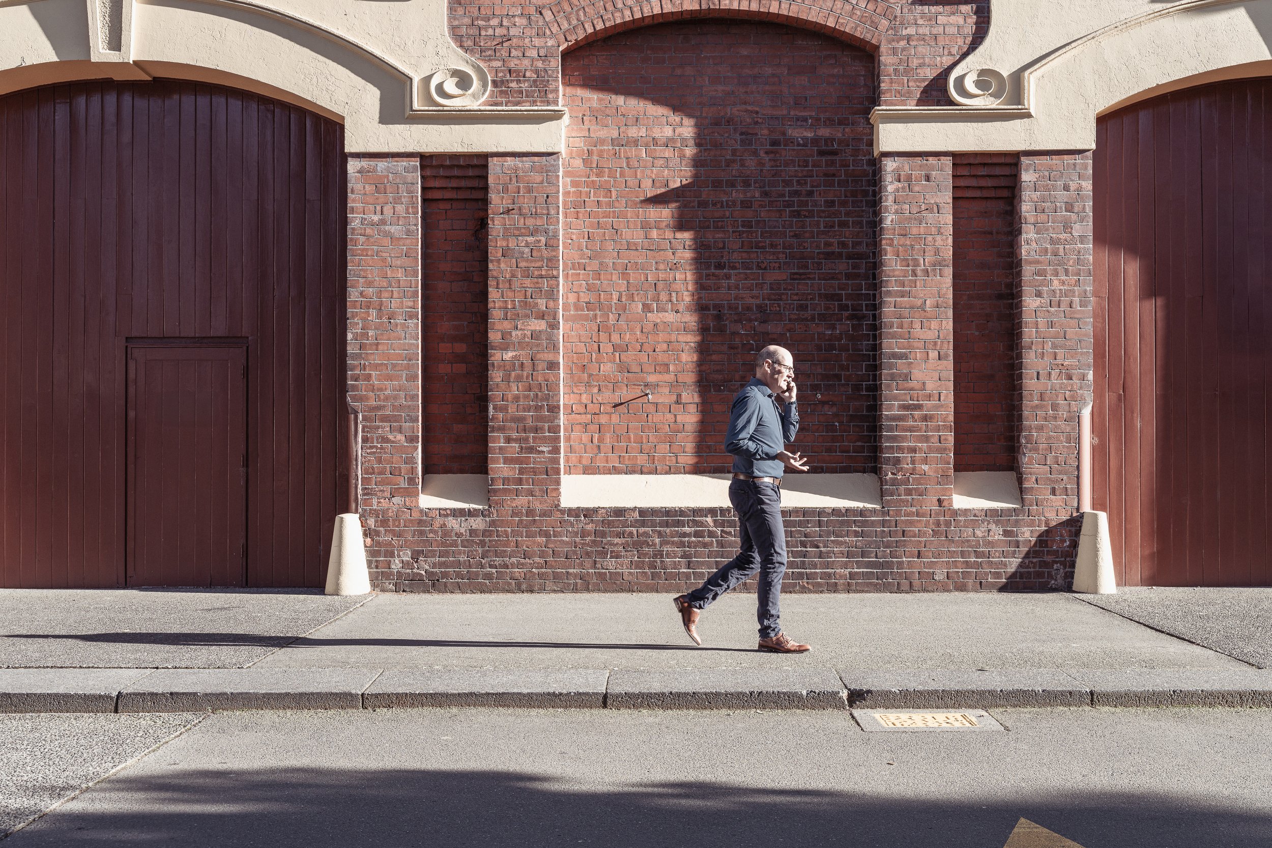 A man walking along a sidewalk talking on his cell phone, with a brick building and closed gates in the background.