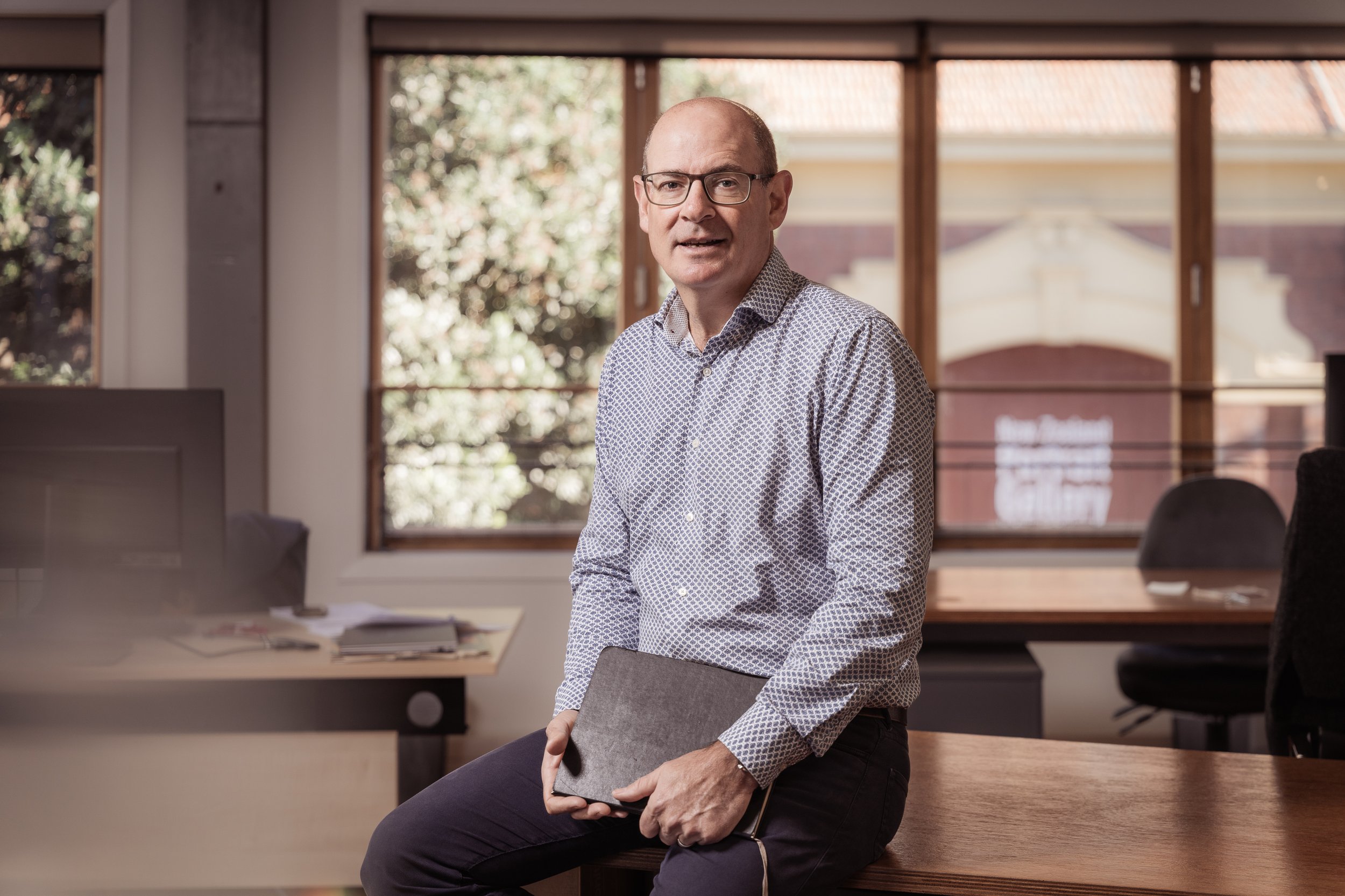 A middle-aged man with glasses and a patterned dress shirt sitting on a wooden table in an office, holding a closed laptop. Behind him are large windows showing trees and a building outside.