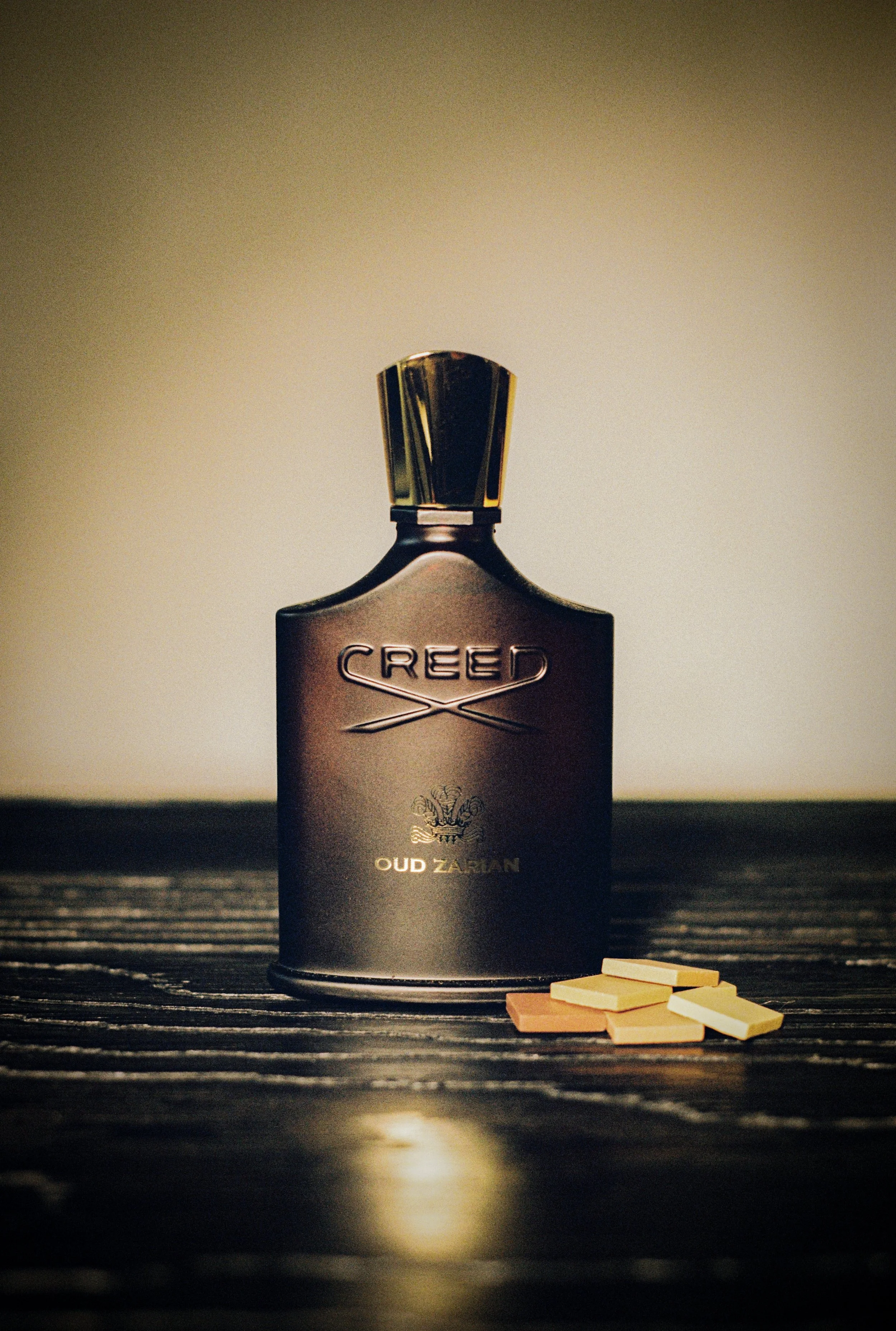 A dark bottle with the label 'CREED' and 'OUD ZARJIAN' is placed on a wooden surface. To the right of the bottle, there are several small rectangular perfume sample cards. The background is neutral beige with focused lighting on the bottle, creating 