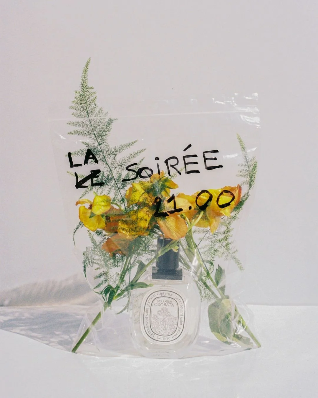 A small bouquet of yellow and orange flowers with green foliage in a clear plastic wrapping with black handwritten text 'LA LE SOIRÉE 21.00' and a white label at the bottom.