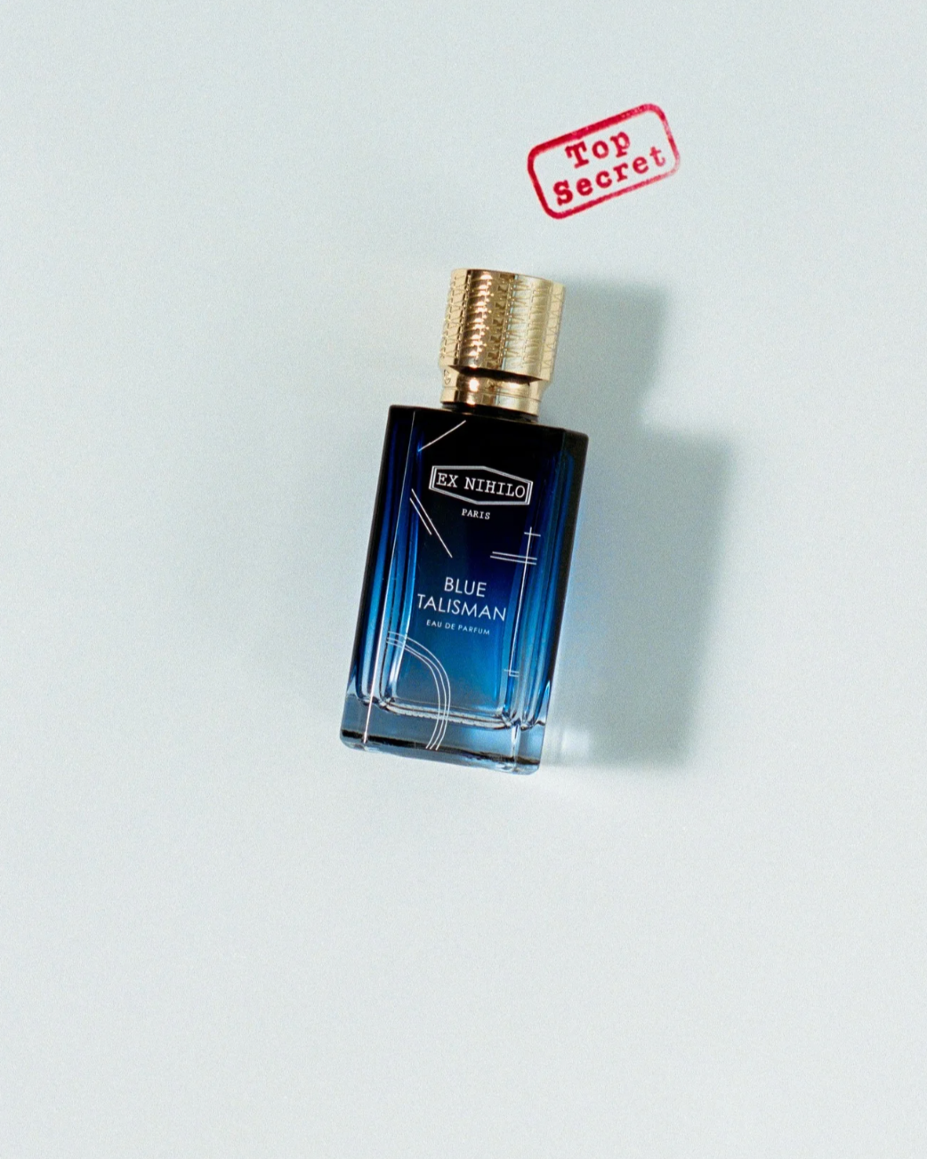 A perfume bottle labeled 'EX NIHILO PARIS BLUE TALISMAN EAU DE PARFUM' with a gold cap, placed on a white surface. A red stamp reads 'Top Secret' in the upper right of the image.