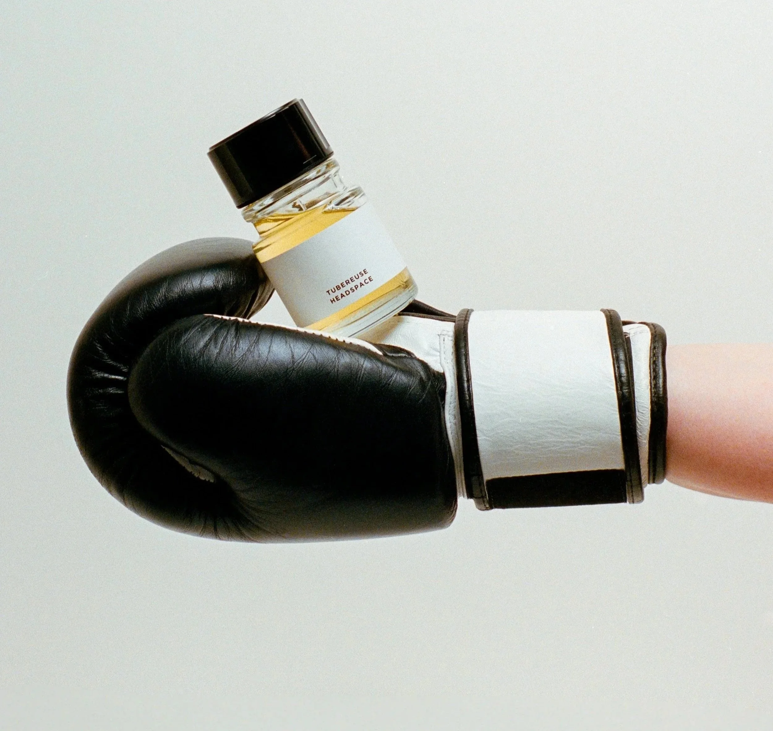 Small glass bottle labeled 'Tubereuse Headspace' being held by a person wearing black gloves, against a plain grey background.