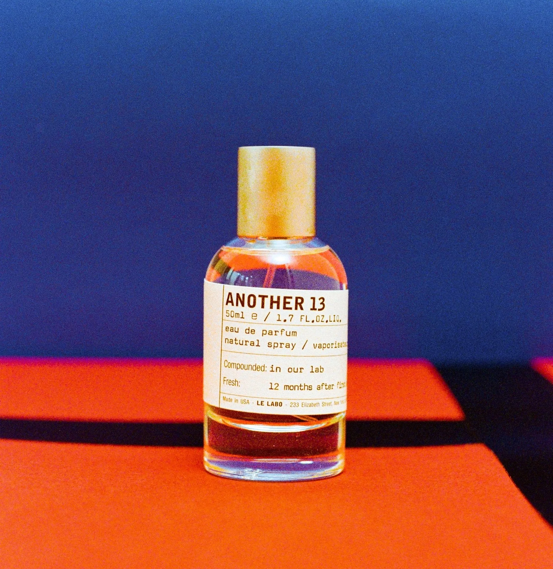 A small perfume bottle labeled 'Another 13' against a blue background, with a blue and orange surface below.