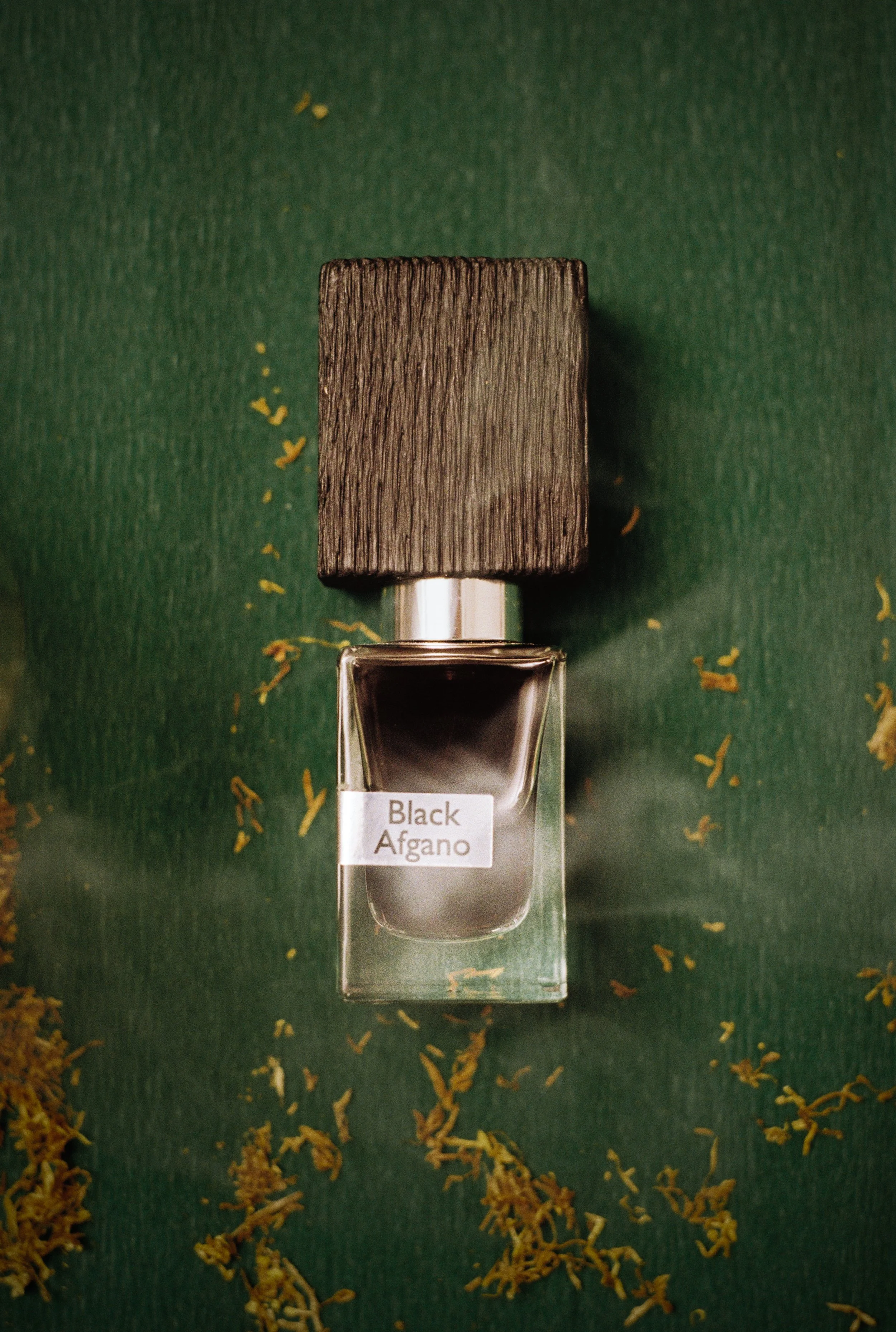 A bottle of Black Affano perfume with a wooden cap, placed on a green surface scattered with dried flower petals.