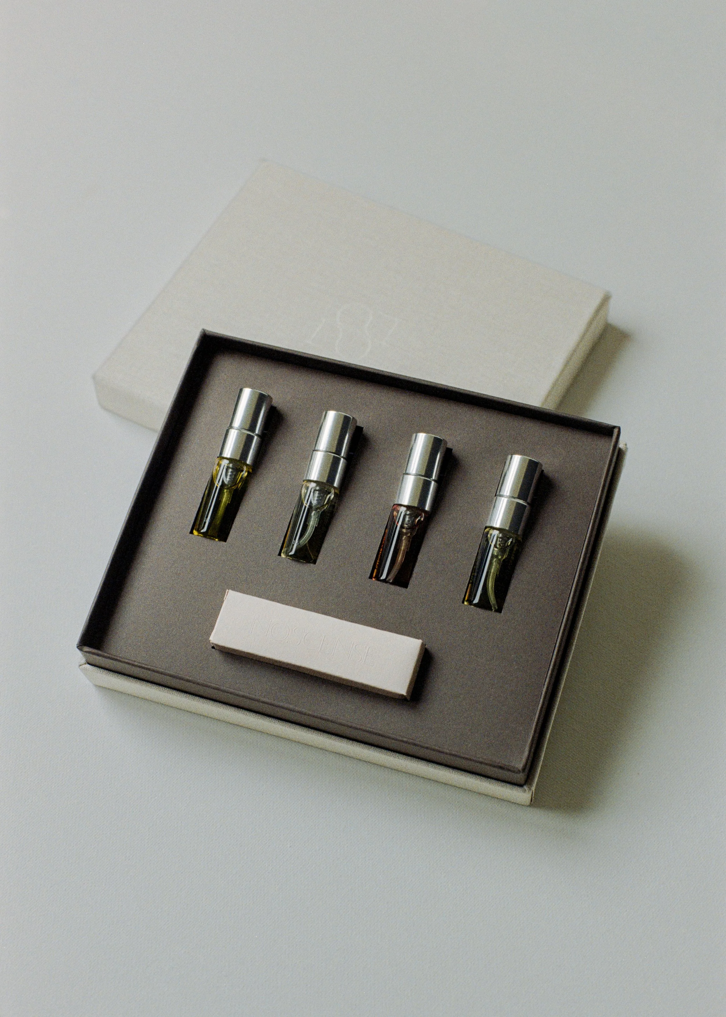 A set of four glass perfume bottles with silver caps inside a gray box with foam padding. The box is placed on a white surface, and its lid is partially visible in the background.
