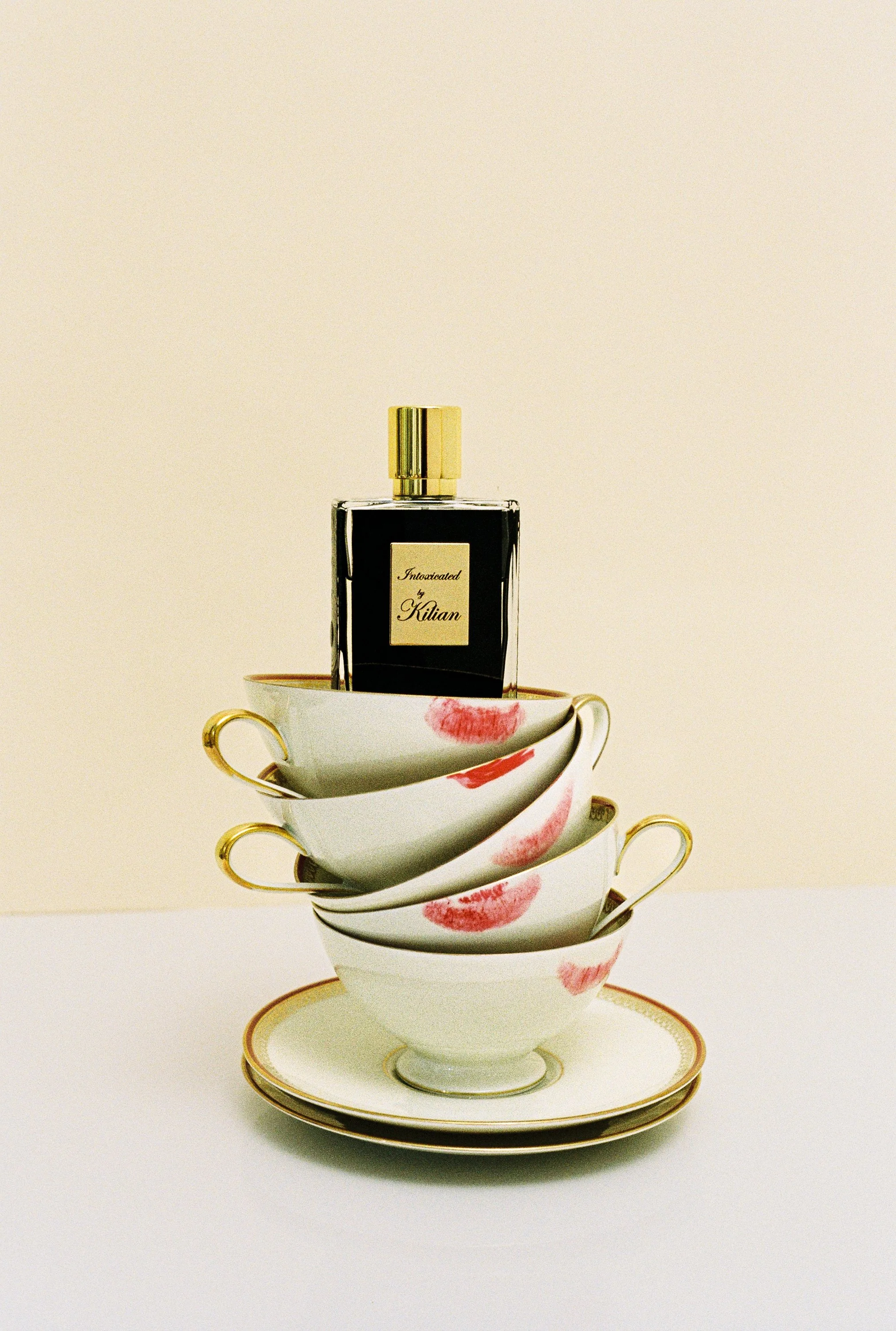 Stacked teacups with pink lipstick kiss marks, topped by a perfume bottle labeled 'Intoxicated by Kitian' on a plain background.
