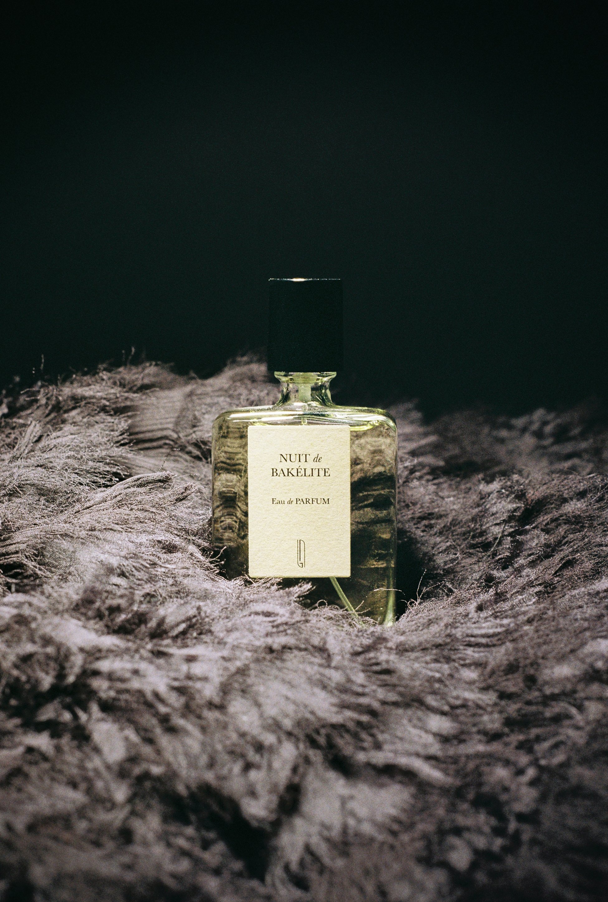 A perfume bottle labeled 'NUIT de BAKÉLITE' resting on a bed of fluffy, light-colored feathers.