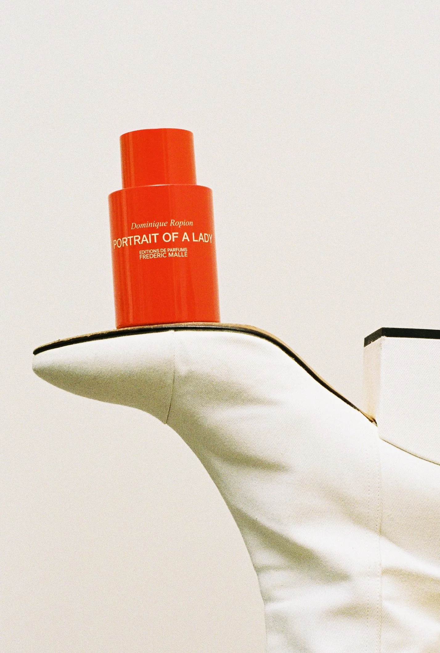 A red cylindrical perfume bottle labeled 'Portrait of a Lady' by Dominique Ropion, placed on a curved white shelf against a plain white wall.