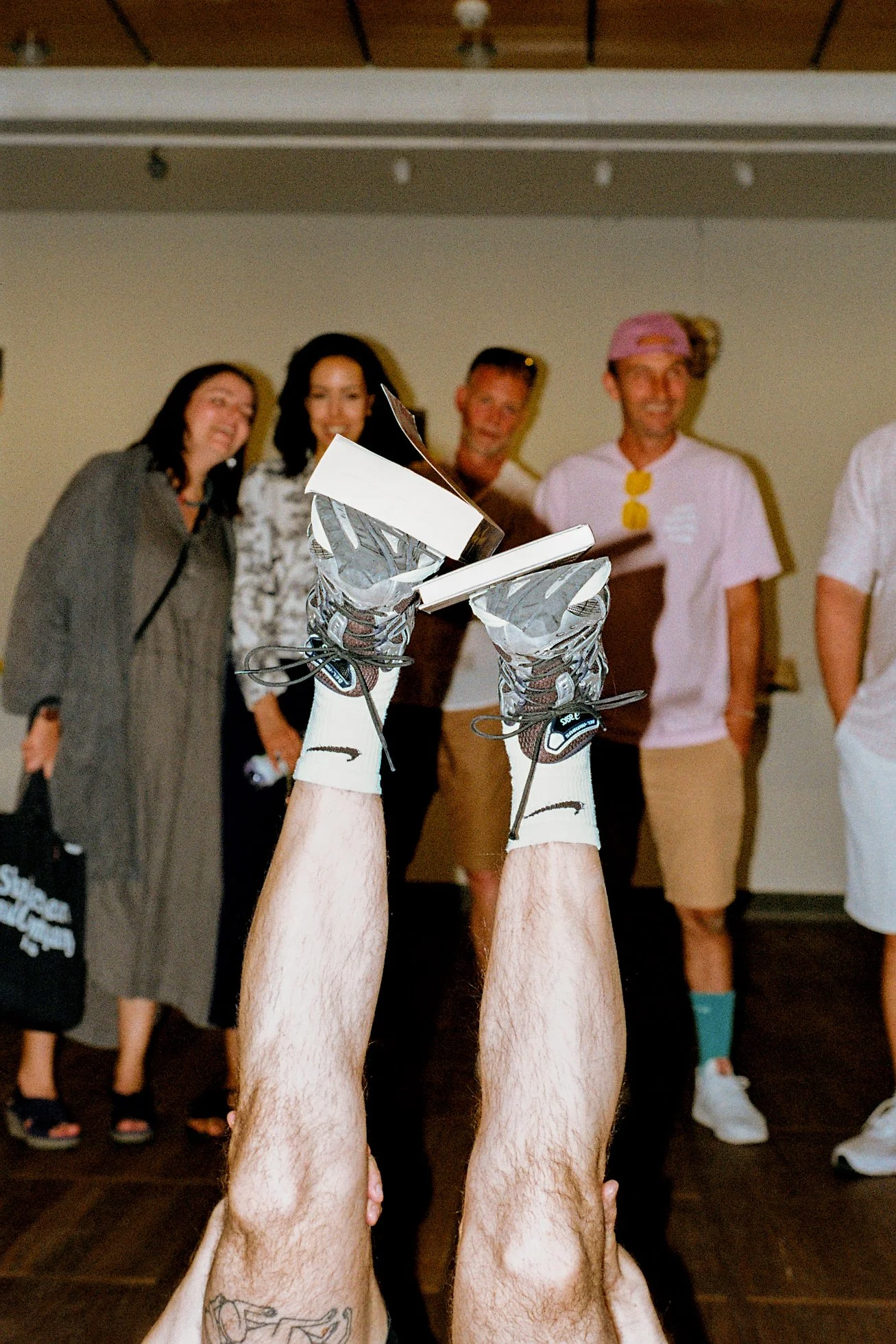 Person lying on their back with legs raised in the air, wearing running shoes, in front of a group of people standing and smiling indoors.