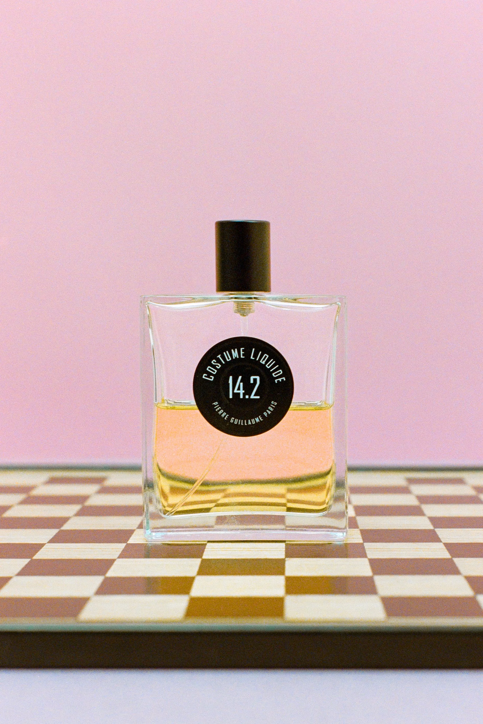 A square glass perfume bottle with a black spray cap, placed on a checkered surface with a pink background. The bottle has a round black label with white text that reads 'Costume Liquide 14.2 Pierre Guillaume Paris'.