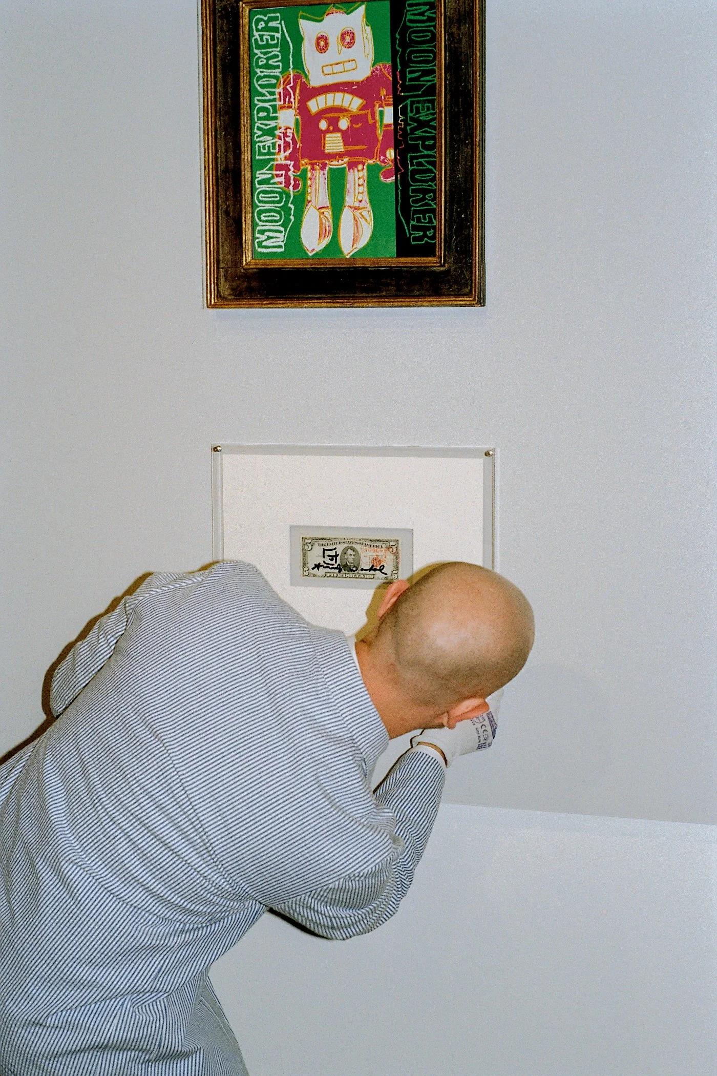 A man with a bald head, wearing a long-sleeved, light blue and white striped shirt, is sitting at a table with his head resting on his hand, looking at a framed picture on the wall. There is a one-dollar bill pinned to the wall below a colorful Frida Kahlo artwork.