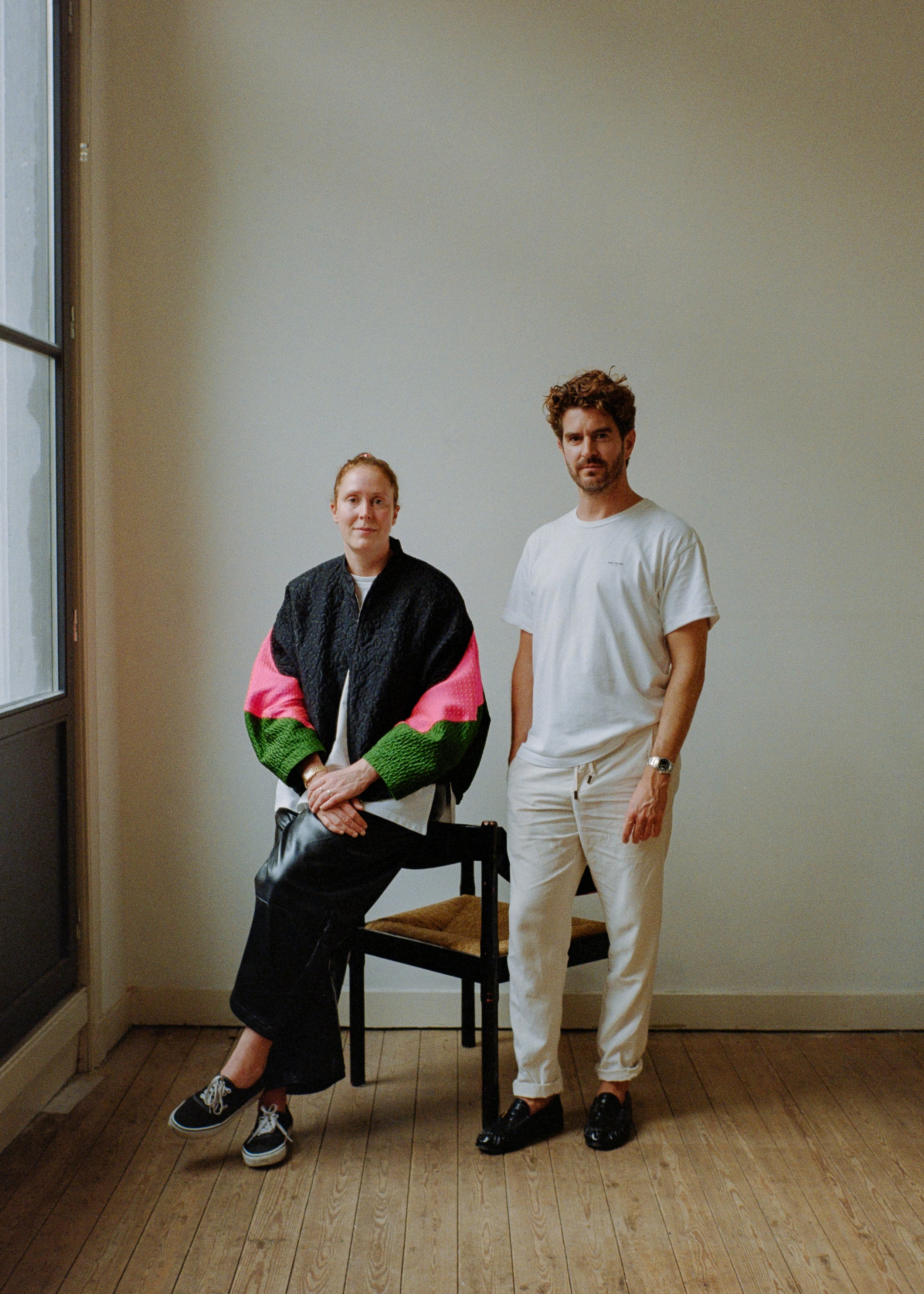 A woman with red hair and a man with dark, curly hair standing in a room with wooden floors and beige walls. The woman is seated on a chair, wearing a colorful oversized jacket and black pants, while the man is standing next to her, dressed in white shirt and pants. They are looking at the camera.