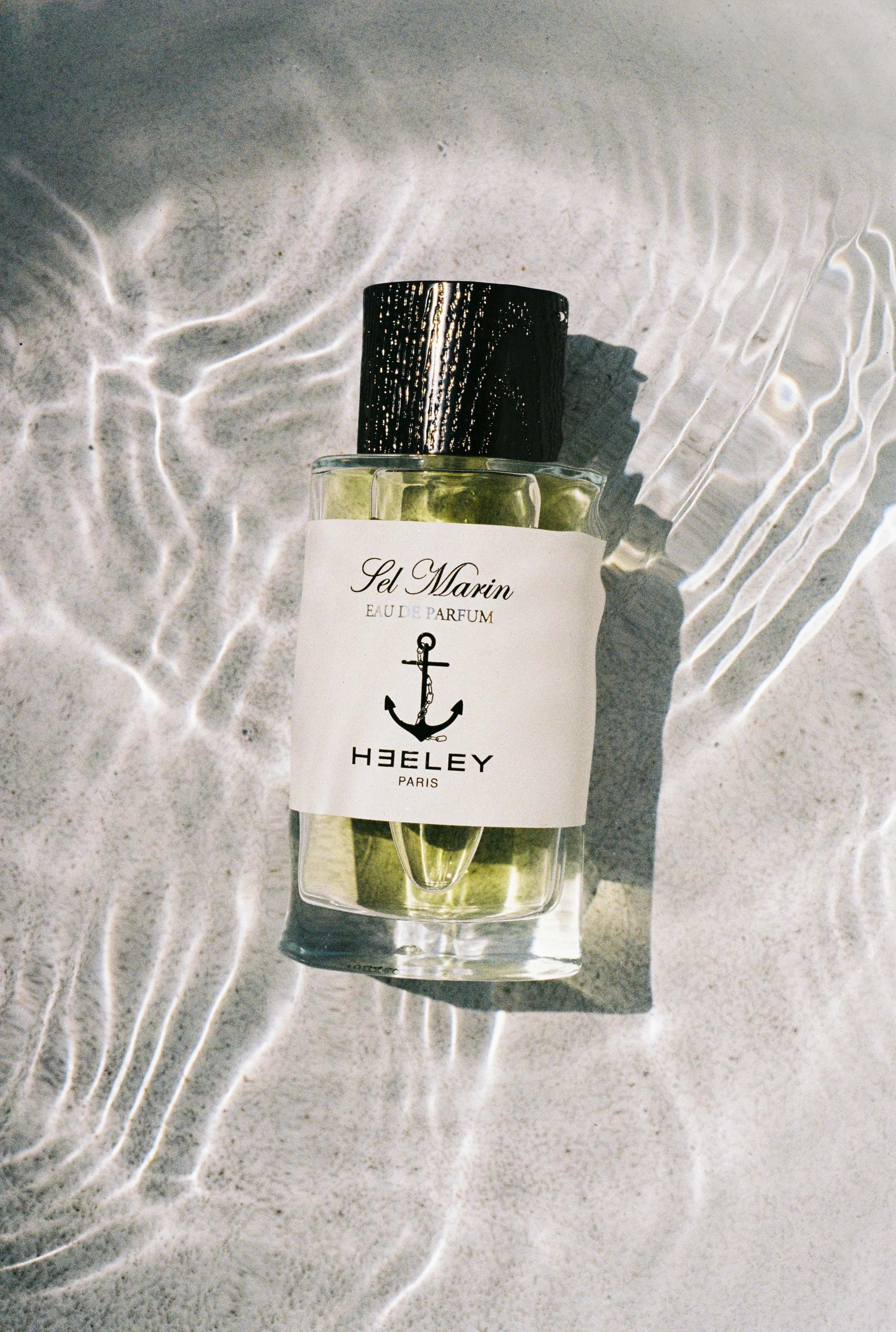 A bottle of Le Marin Eau de Parfum with a black cap, placed on textured sand with sunlight creating reflections and shadows.