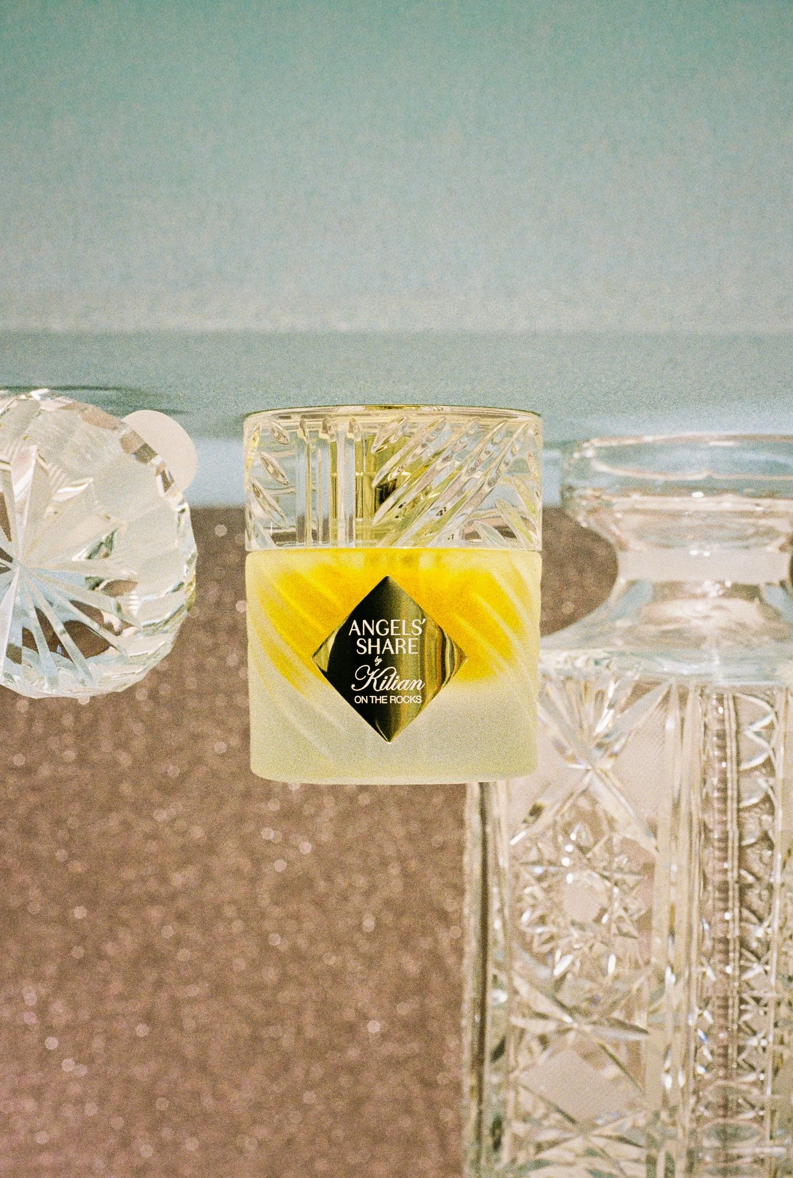 A clear glass container with a diamond-shaped pattern and a yellow label that reads 'Angels Share by Kilian on the Rocks', placed on a reflective table beside decorative glass objects.