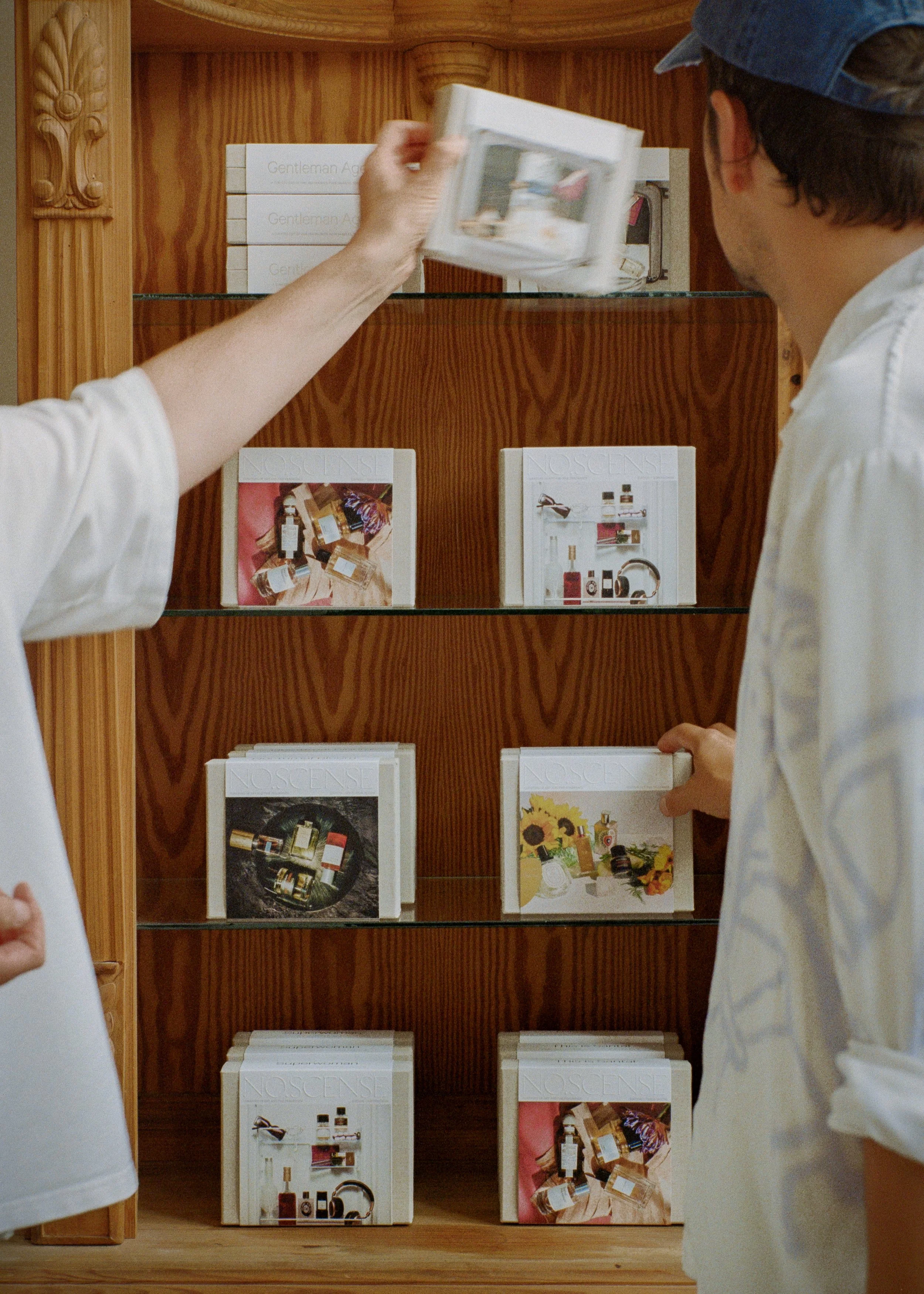 Two people are browsing a wooden display shelf with skincare products and books. One person is reaching out to pick an item, while the other is holding a booklet. The products and booklets are neatly arranged on the shelf.