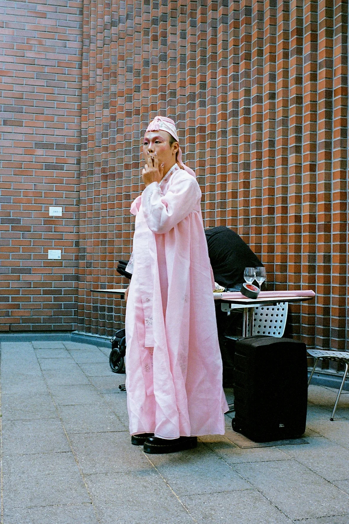 A person dressed in a long pink gown and a pink hat stands against a brick wall, touching their lips with fingers, with a table and chairs behind them and a person bent over at the table.