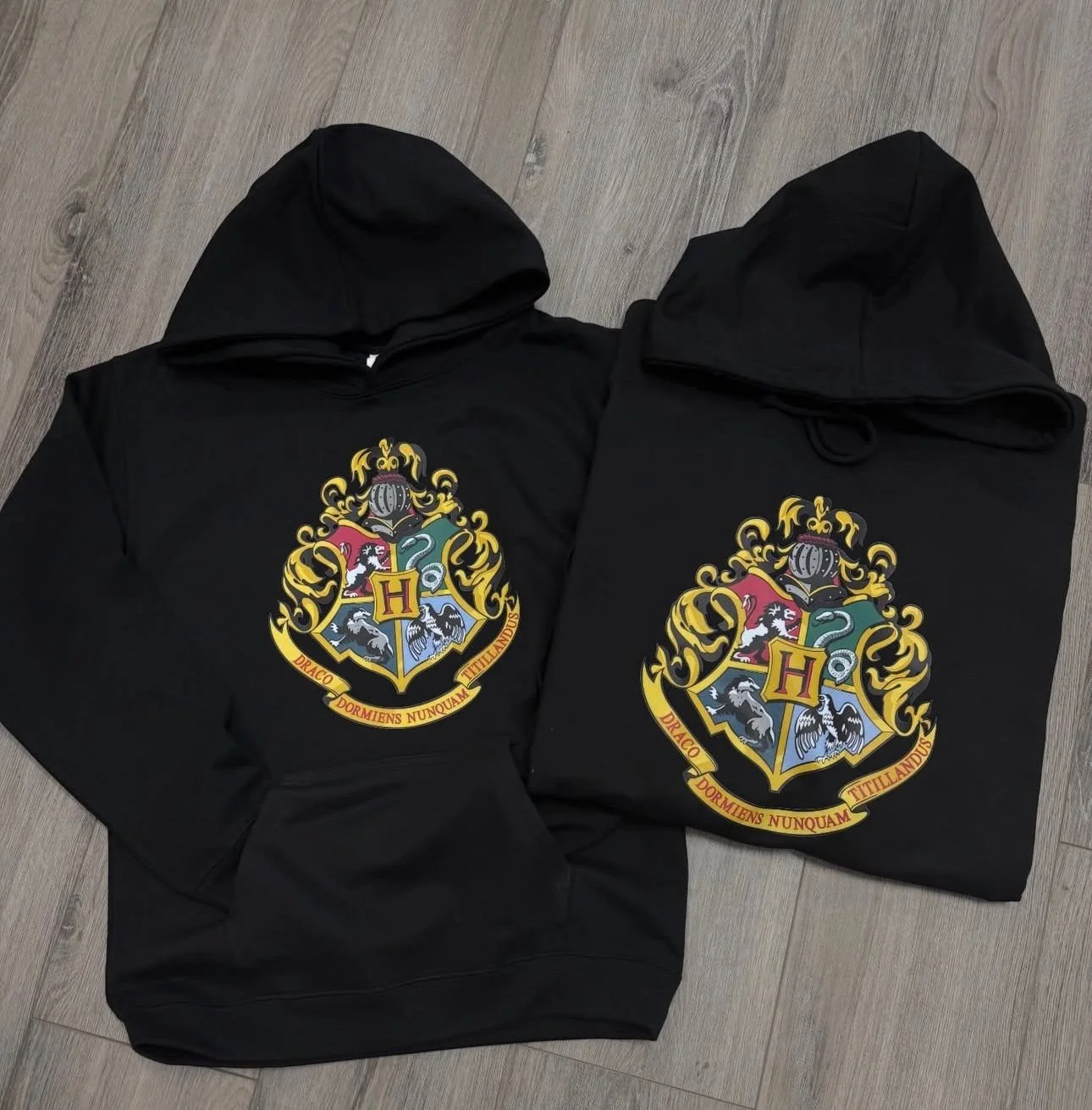 Two black hoodies with the Hogwarts crest printed on the front, laid out on a wooden floor.