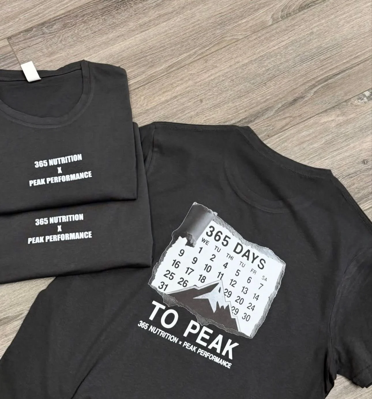 Three black T-shirts with white text and graphics, laid out on a wooden floor. Two shirts say '365 NUTRITION X PEAK PERFORMANCE,' and one shirt features a calendar graphic with mountain scenery and the text '365 DAYS TO PEAK'.