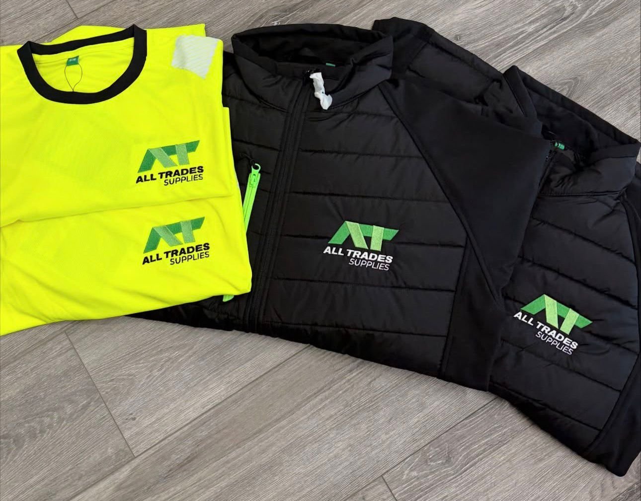 Bright yellow safety vest and black insulated jacket, both with the logo 'All Trades Supplies' printed on the front.