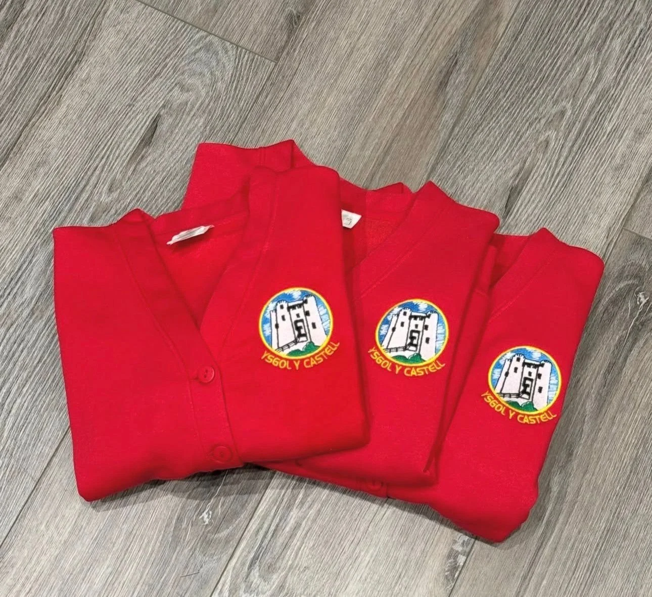 Three folded red polo shirts with an embroidered crest of Ysgol Y Castell, a castle illustration and the school name, placed on gray wooden floor.