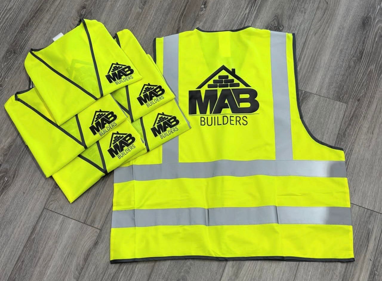 High visibility yellow safety vests with black and gray reflective stripes, labeled "MAB BUILDERS" with a house and brick wall logo, laid out on a wooden floor.
