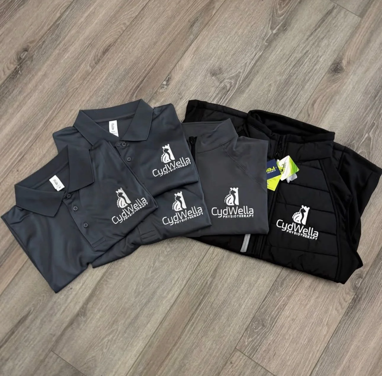Three polo shirts and a pair of shorts with 'CydWella Physiotherapy' logos laid on a wooden floor.