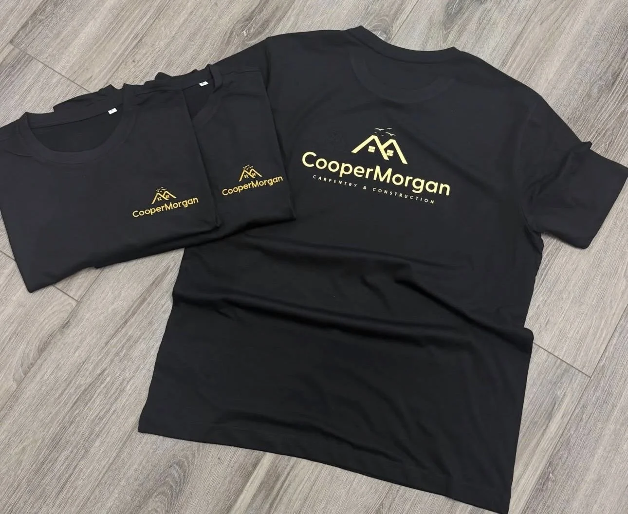 Black T-shirts with yellow logo and text 'Cooper Morgan' and 'Carpentry & Construction' printed on front, laid flat on a wooden floor.