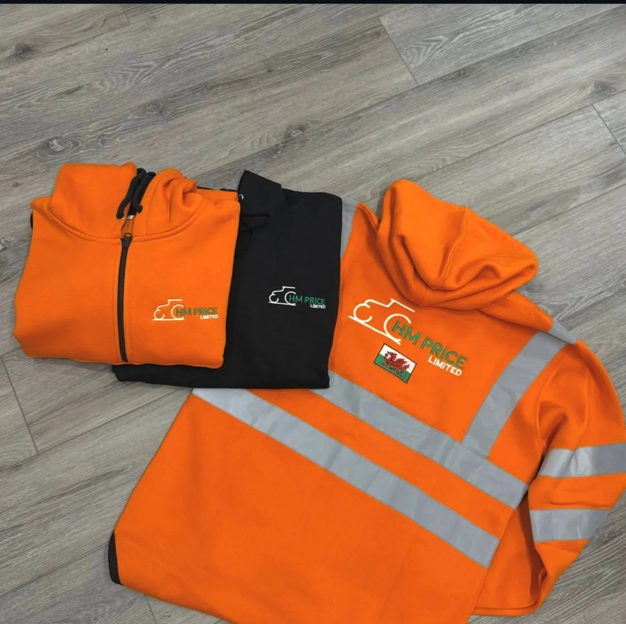 Three pieces of clothing with 'HM PRICE LIMITED' logo on a light gray wooden floor: a folded orange zip-up hoodie, a folded black hoodie, and a high-visibility orange safety jacket with reflective stripes and logo.