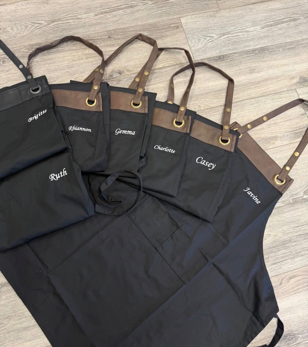 Set of black apron and matching kitchen towels with brown straps and gold accents, each towel personalized with a white embroidered name, laid on a light wood floor.