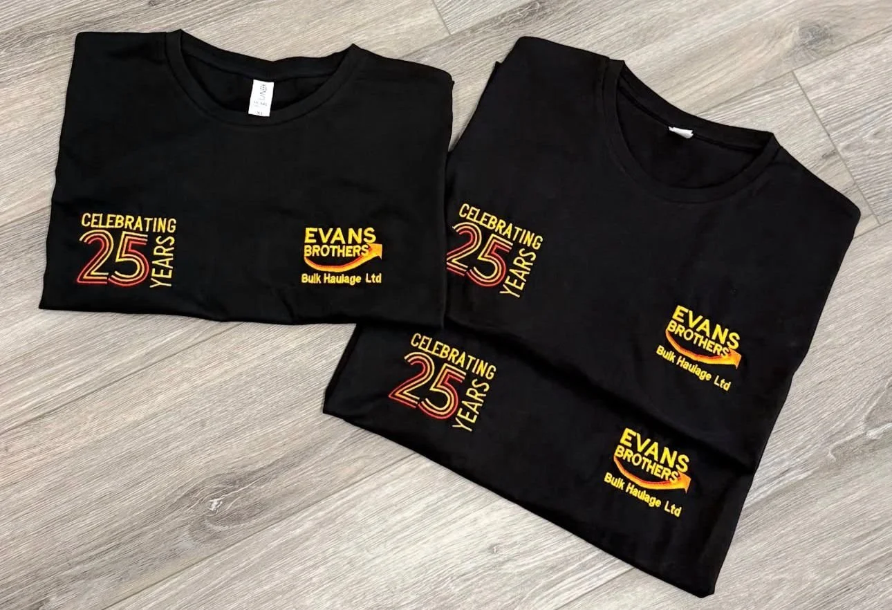 Two black T-shirts with yellow and red text celebrating 25 years of Evans Brothers Bulk Haulage Ltd, laid on a gray wooden surface.