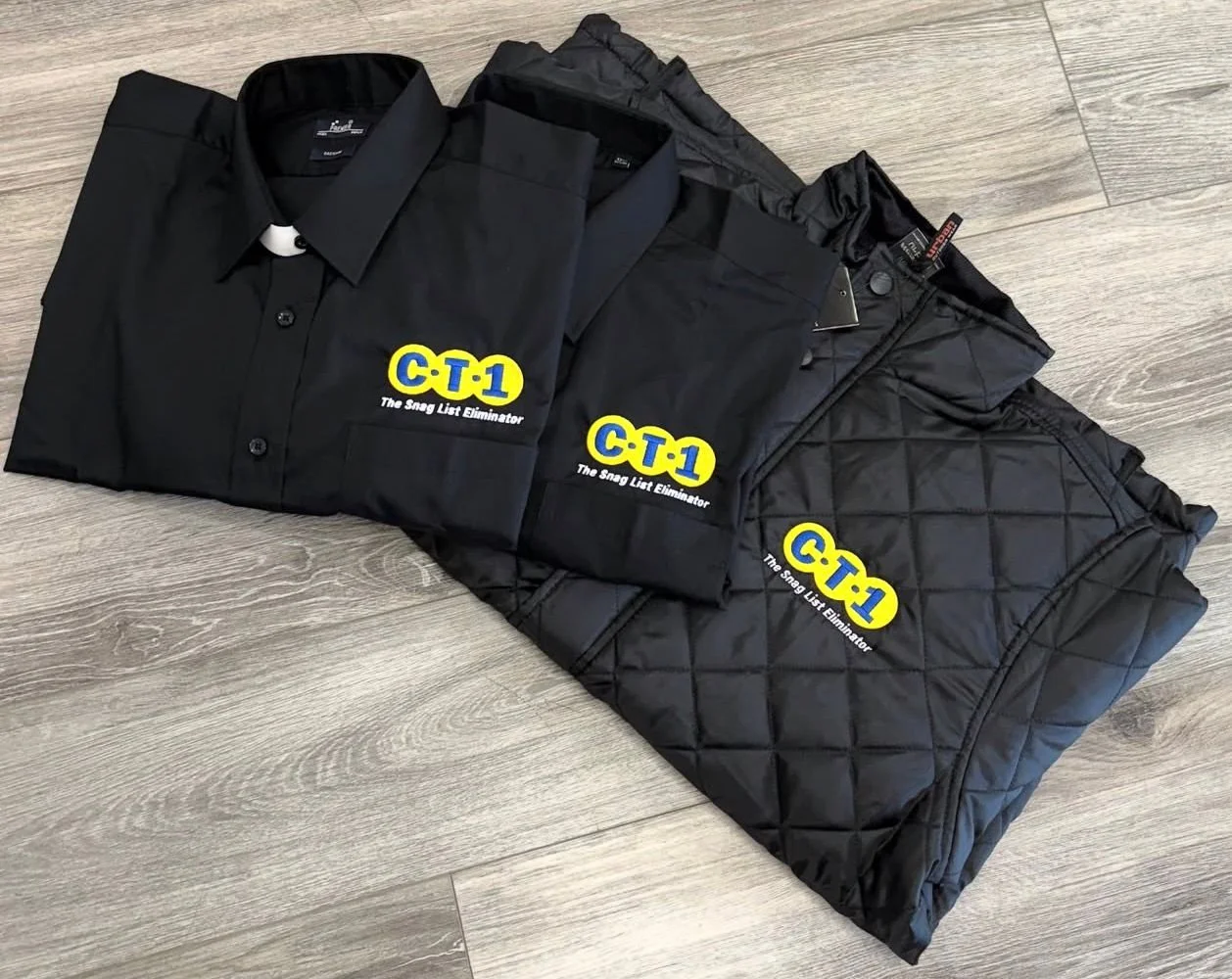 Three black jackets with logos reading "C-T-1 The Snag List Eliminator" laid out on a wooden floor.