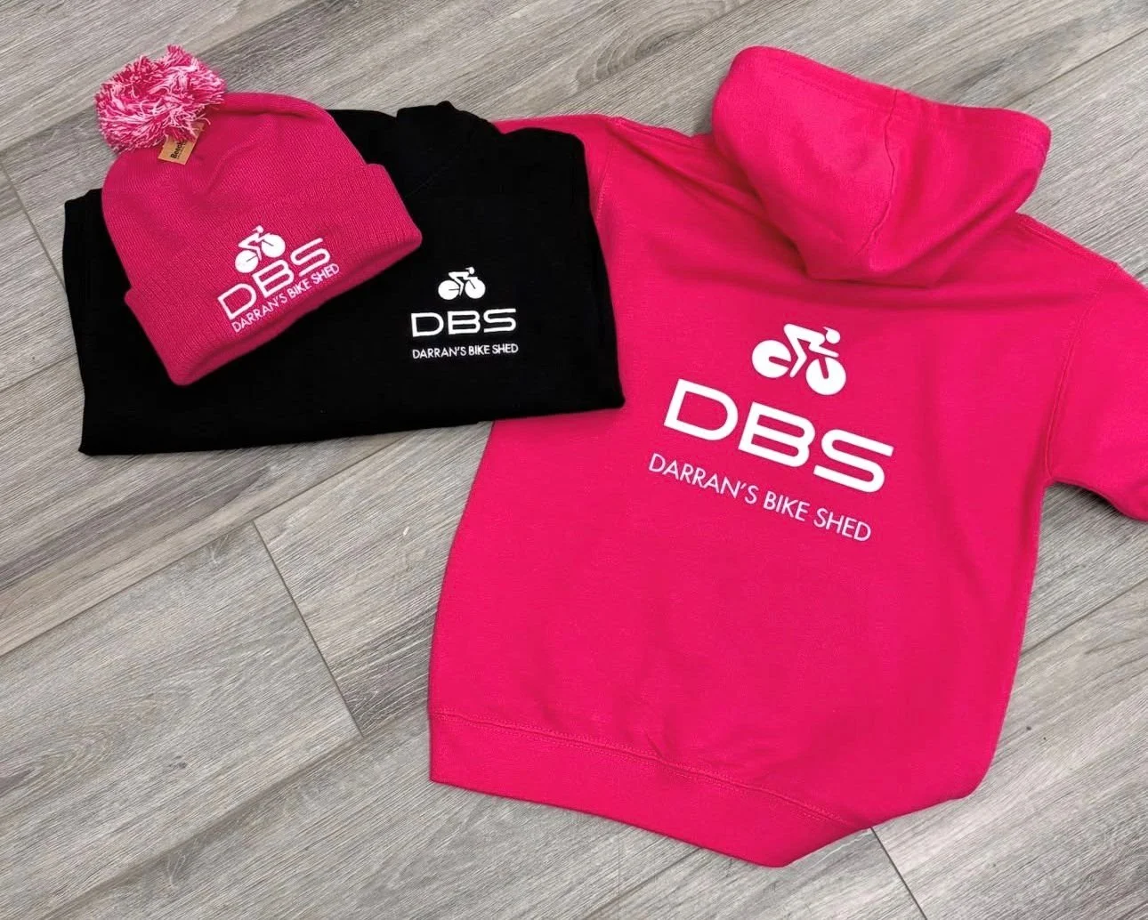Pink and black sweatshirts and a pink beanie hat with logo and text for Darran's Bike Shed on a wood floor.