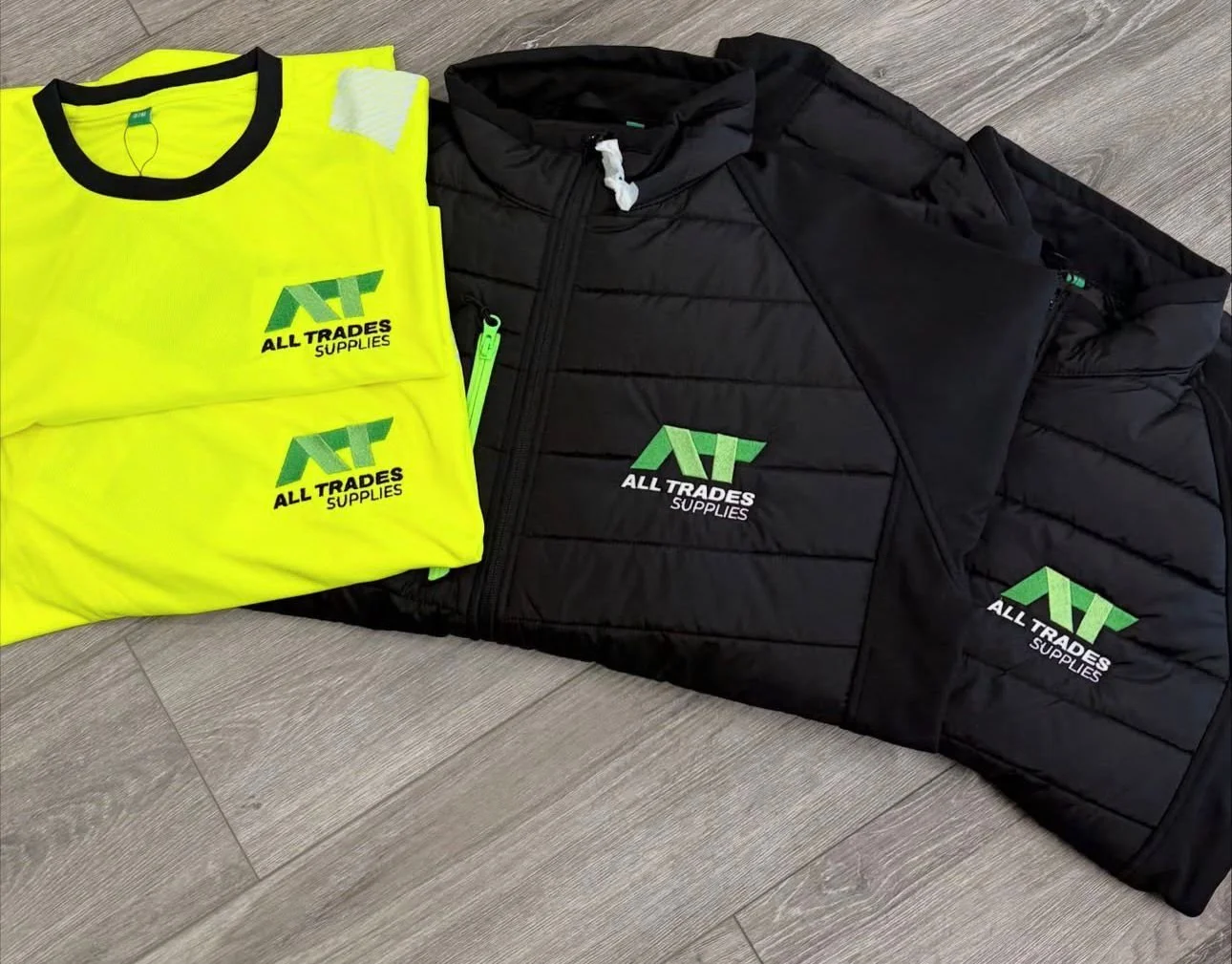 Two yellow high-visibility shirts and two black jackets with the logo "All Trades Supplies" laid out on a wooden floor.