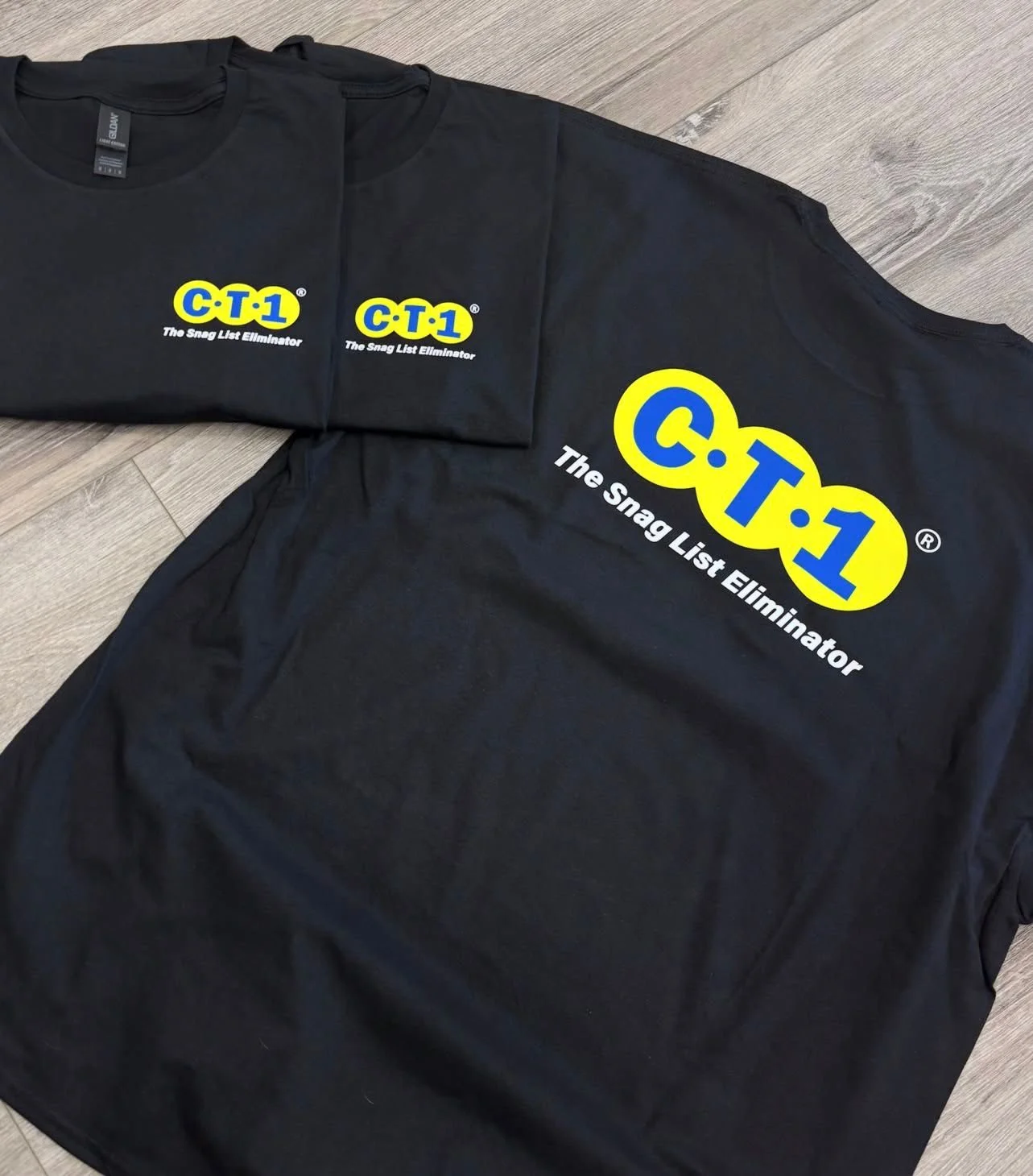 Black t-shirts with the logo 'C.T-1' and the tagline 'The Snag List Eliminator' printed on the front, laid out on a wooden surface.