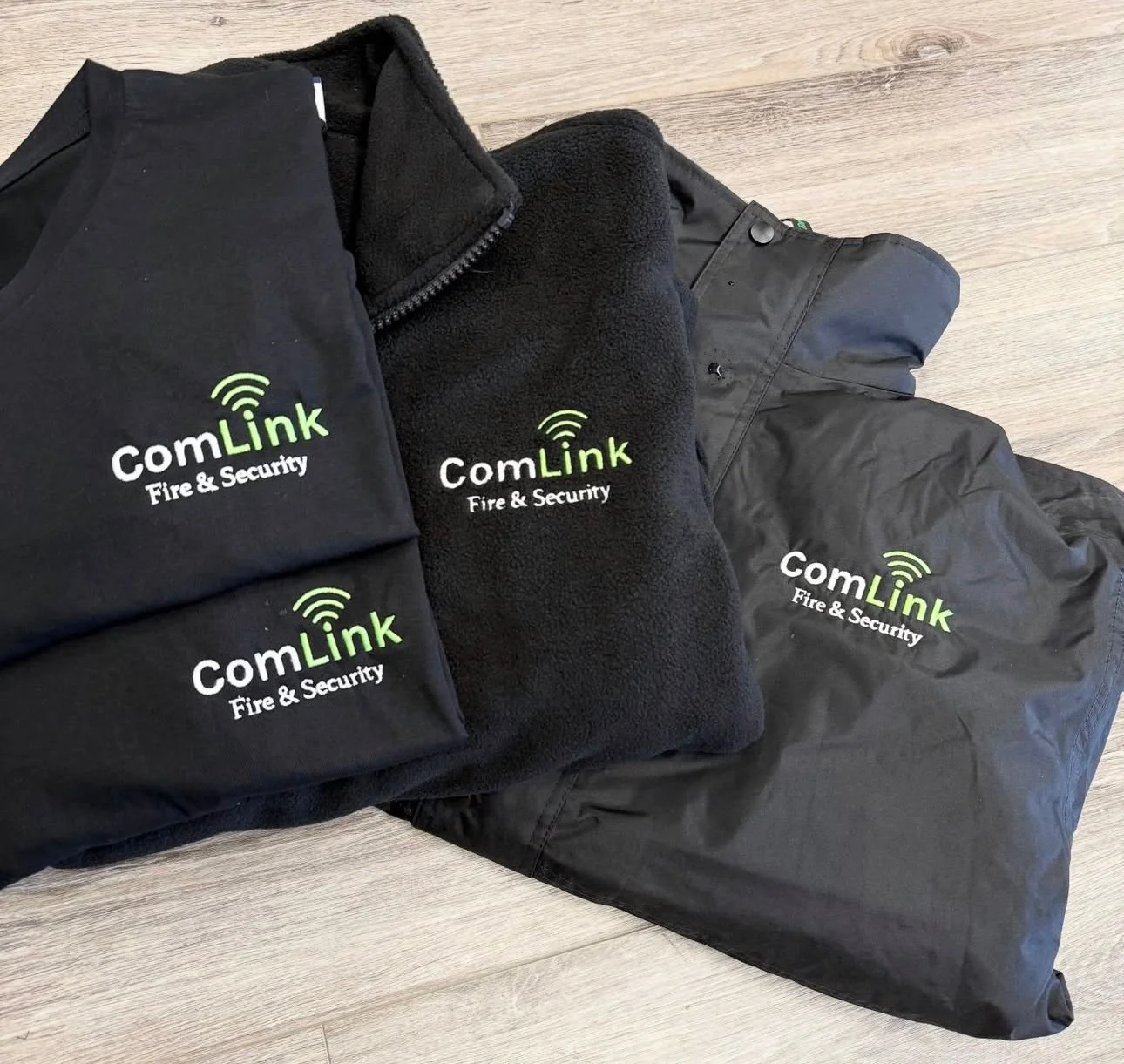 Three black jackets and a black bag, all with the logo and text 'ComLink Fire & Security' in green and white, laid out on a light gray wooden floor.