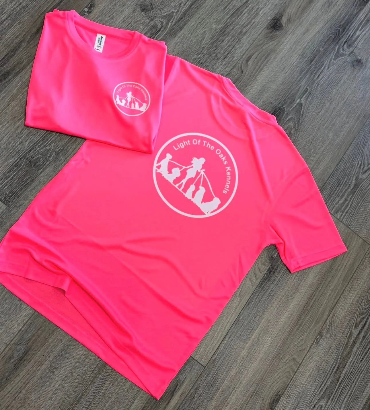 Bright pink t-shirt and towel with a white circular logo featuring a person hiking and text that reads "Light Of The Oaks Kemnels" on a gray wood floor.