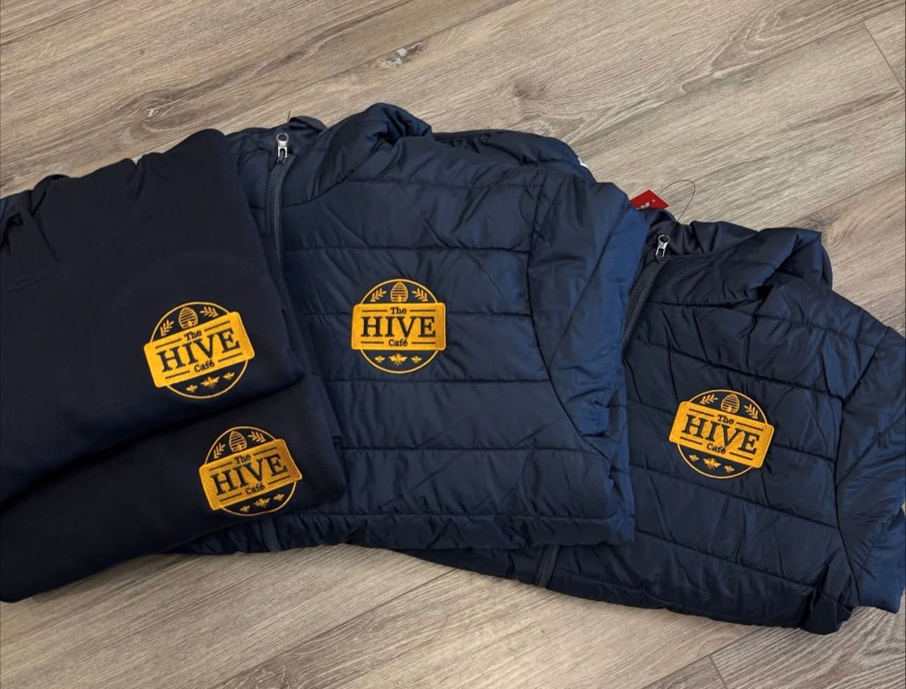 Three navy blue jackets with yellow embroidered patches on a wooden floor. The patches read 'The HIVE Cafe' with a hive and bee design.