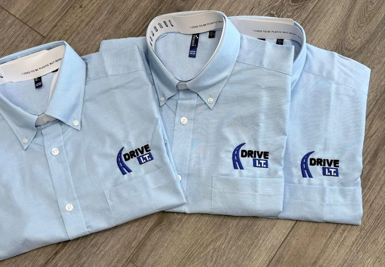 Three light blue button-down shirts with embroidered company logos reading "Drive I.T." on the left chest, laid out on a wooden surface.